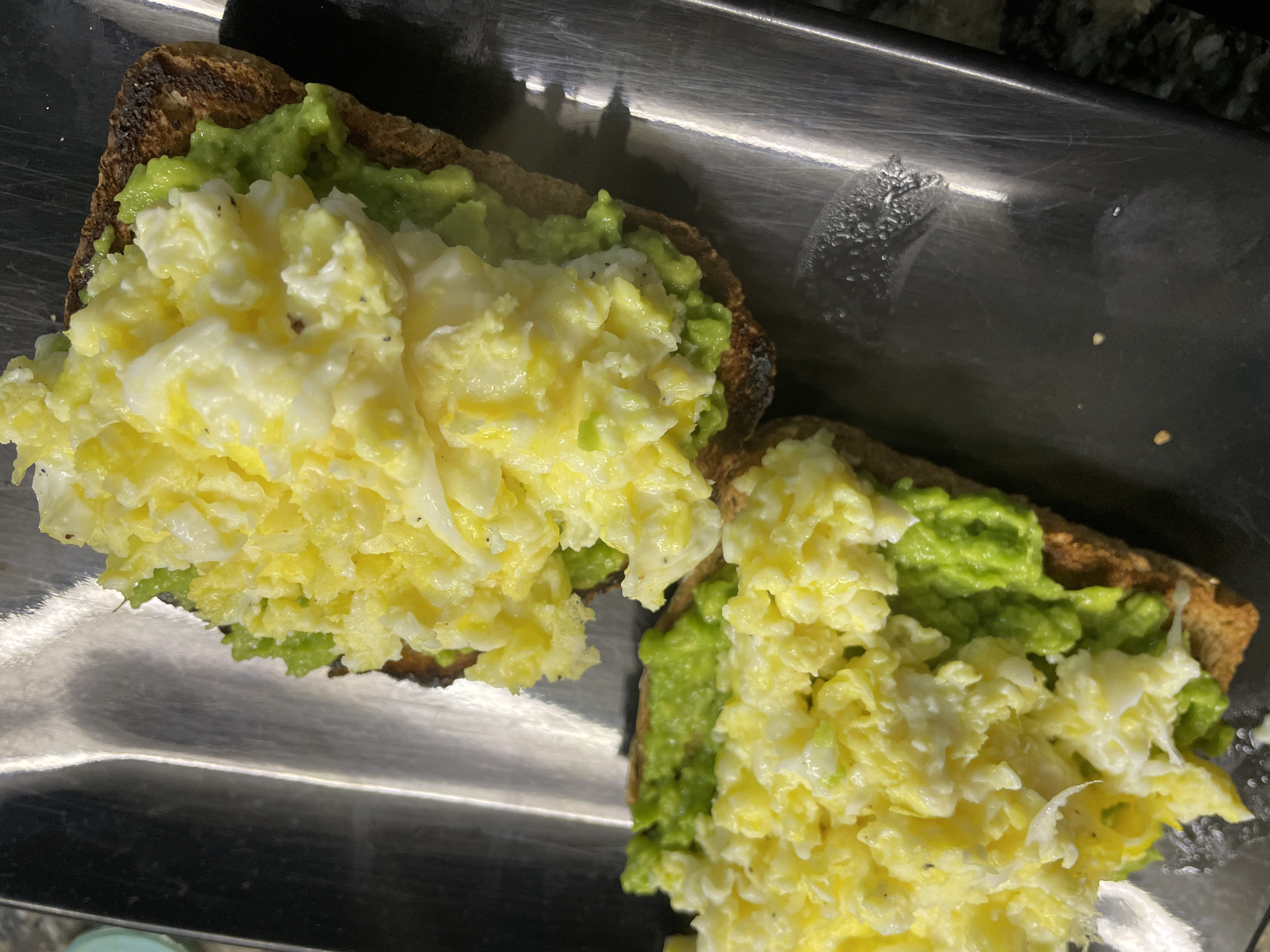 An overhead shot displays two pieces of avocado toast, each topped with a generous portion of scrambled eggs, resting on a dark, reflective rectangular tray or serving dish. The toast appears to be whole wheat or a similar dark bread, with some visible charring on the edges, particularly on the left piece. A layer of mashed green avocado is spread over each slice of toast, acting as a base for the eggs. The scrambled eggs are light yellow and white, with a soft, fluffy texture, and a few dark specks, likely ground pepper, are visible.
The serving surface is dark, possibly metal, and shows signs of use, including scratches, smudges, and some areas with what appears to be condensation or water droplets, especially visible in the upper right and bottom right sections of the tray. Reflections of overhead lighting or other objects are cast upon the shiny surface of the tray. The food is neatly arranged, suggesting it is prepared for a meal, likely breakfast or brunch given the components. No people are visible, and there is no discernible text in the image. The provided context indicates the location is Buenos Aires, Argentina.