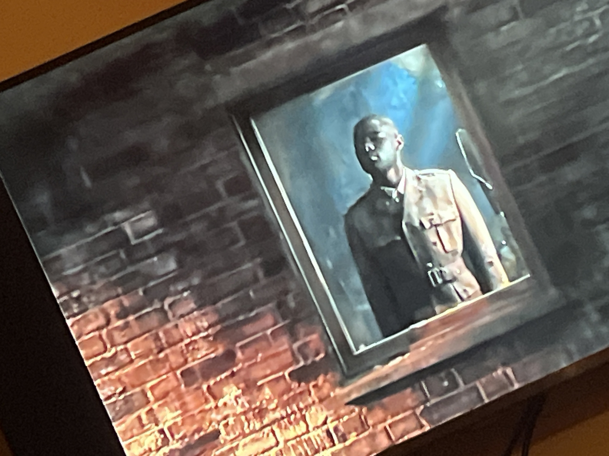 The image displays a framed portrait of a Black man wearing a light-colored military-style uniform with a belt. The portrait is set against a brick wall, which appears to be illuminated from below with a warm, reddish light, suggesting a fire or intense heat source. The portrait itself has a painted background that transitions from dark to a vibrant blue at the top. The overall impression is a staged or artistic representation rather than a candid photograph. The location is Gura Top, Nigeria.