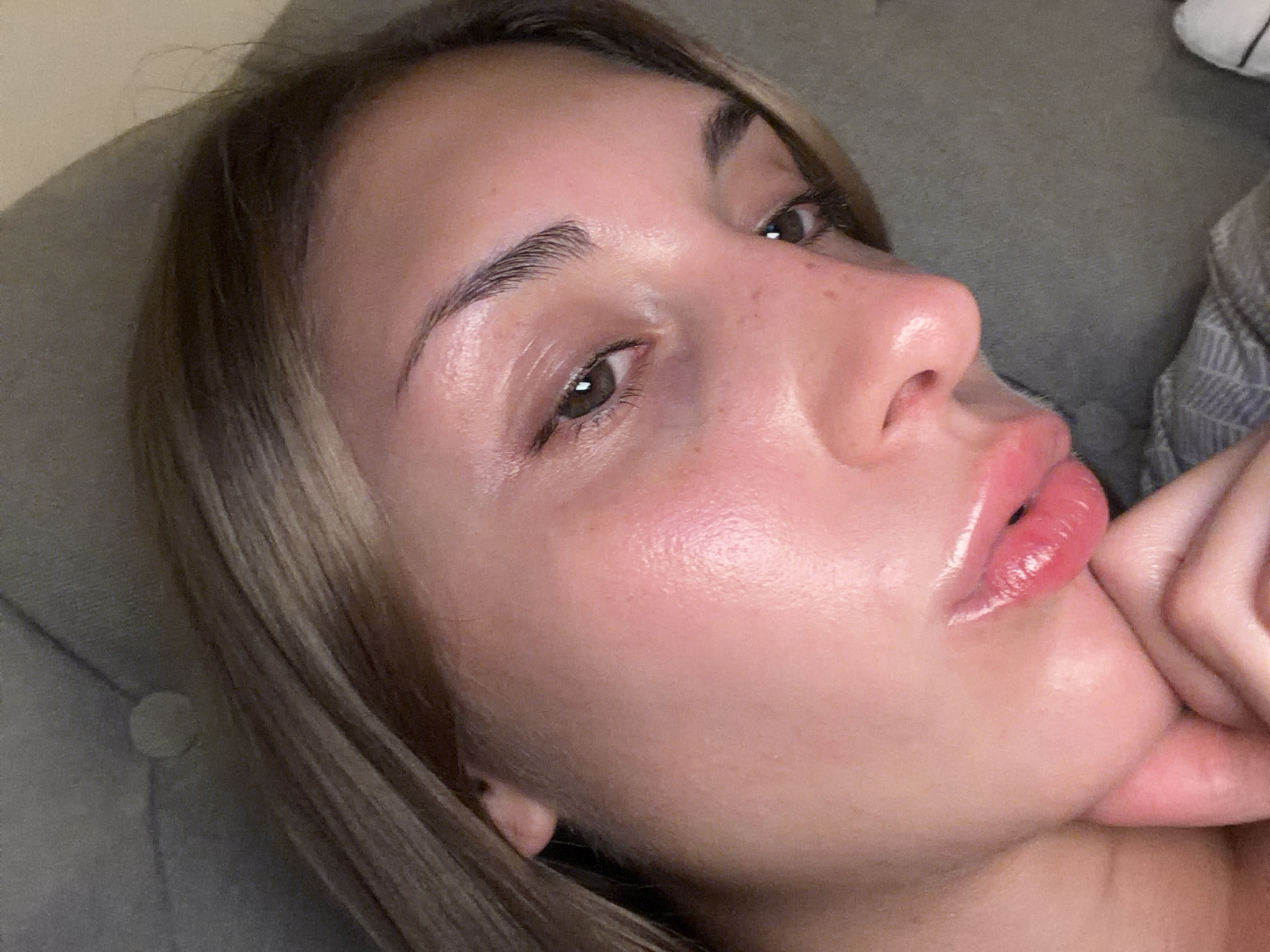A close-up, indoor shot captures the face of a young woman resting on a gray upholstered surface. Her head is tilted to the right, with her hair, a blend of brown and blonde, falling across her forehead and partially obscuring the background. The woman's skin appears luminous and slightly flushed, with a noticeable sheen. Her eyes are open and directed downwards, with dark lashes and prominent eyebrows. The lighting highlights the contours of her nose and lips, which are glossed and slightly parted. Her hand is visible in the foreground, resting near her chin. The overall impression is one of relaxed intimacy within a domestic setting.