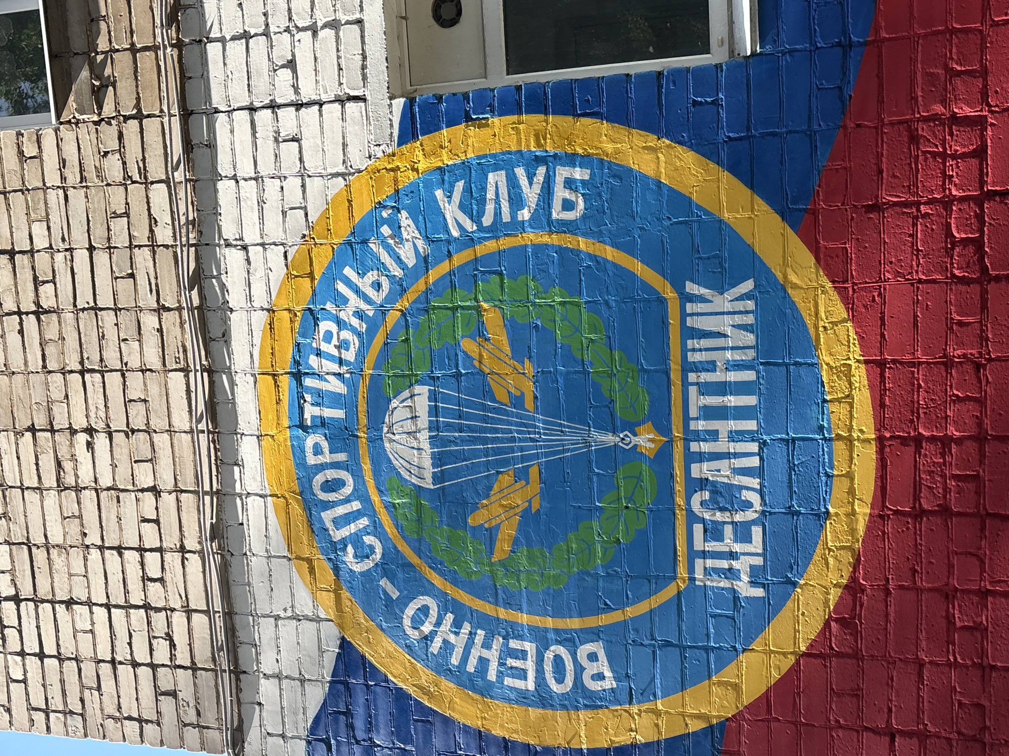 The image displays a close-up of a mural painted on a brick building. The mural features a circular emblem with text and imagery related to a sports club. The emblem is predominantly blue with a yellow border and contains symbols of parachutes and laurel wreaths, suggesting a connection to military or athletic endeavors. White text in Cyrillic script encircles the emblem. On the left side of the emblem, the text reads "СПОРТИВНЫЙ КЛУБ" (SPORTS CLUB), and on the right, it reads "ДЕСАНТНИК" (DESANTNIK, meaning paratrooper or airborne troop). The bottom part of the emblem has the text "ВОЕННО" (VOENNO, meaning military).
The building's exterior is made of light-colored bricks, with some sections painted white and others red, forming a diagonal pattern behind the blue emblem. A window with a white frame is partially visible at the top of the image. The texture of the brickwork is evident through the paint. The lighting suggests it is daytime, with sunlight casting shadows on the textured surface of the bricks and mural. The overall impression is a depiction of a sports club, likely with a military or airborne focus, as indicated by the imagery and text. The mural is located on a building in Chaikovsky, Russia.