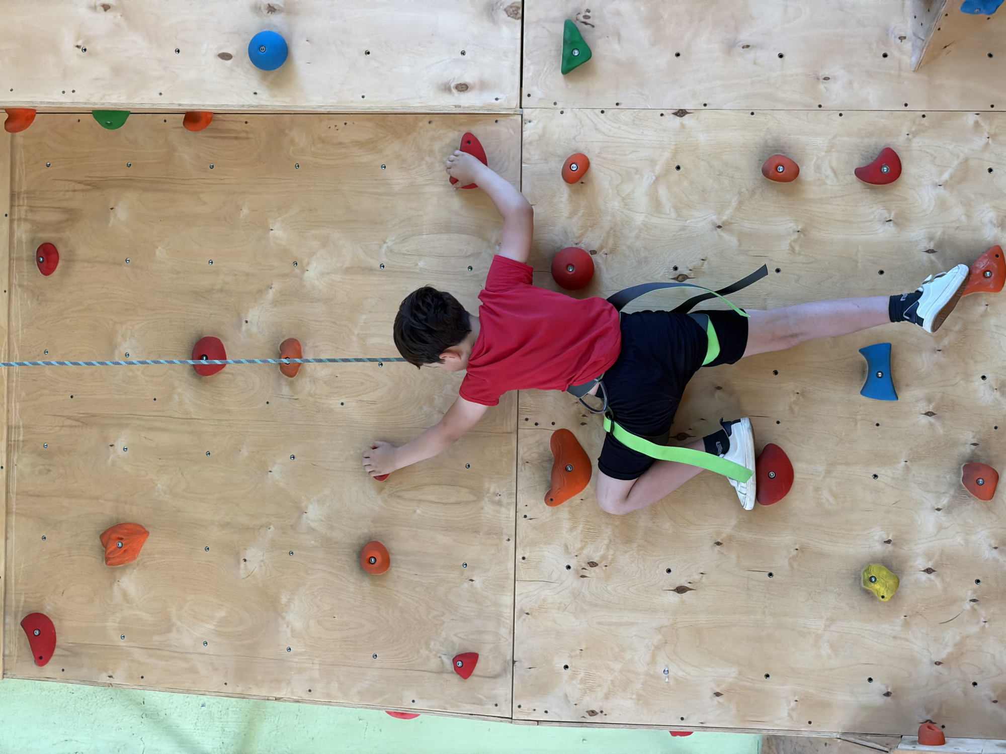 A young boy in a red t-shirt and black shorts is climbing a wooden indoor climbing wall with brightly colored holds. He is wearing a climbing harness with neon green leg loops. The climbing wall appears to be constructed of plywood panels. A grey rope stretches horizontally across the lower section of the wall. The location of the image is not specified as being located in Izhevsk, Russia.