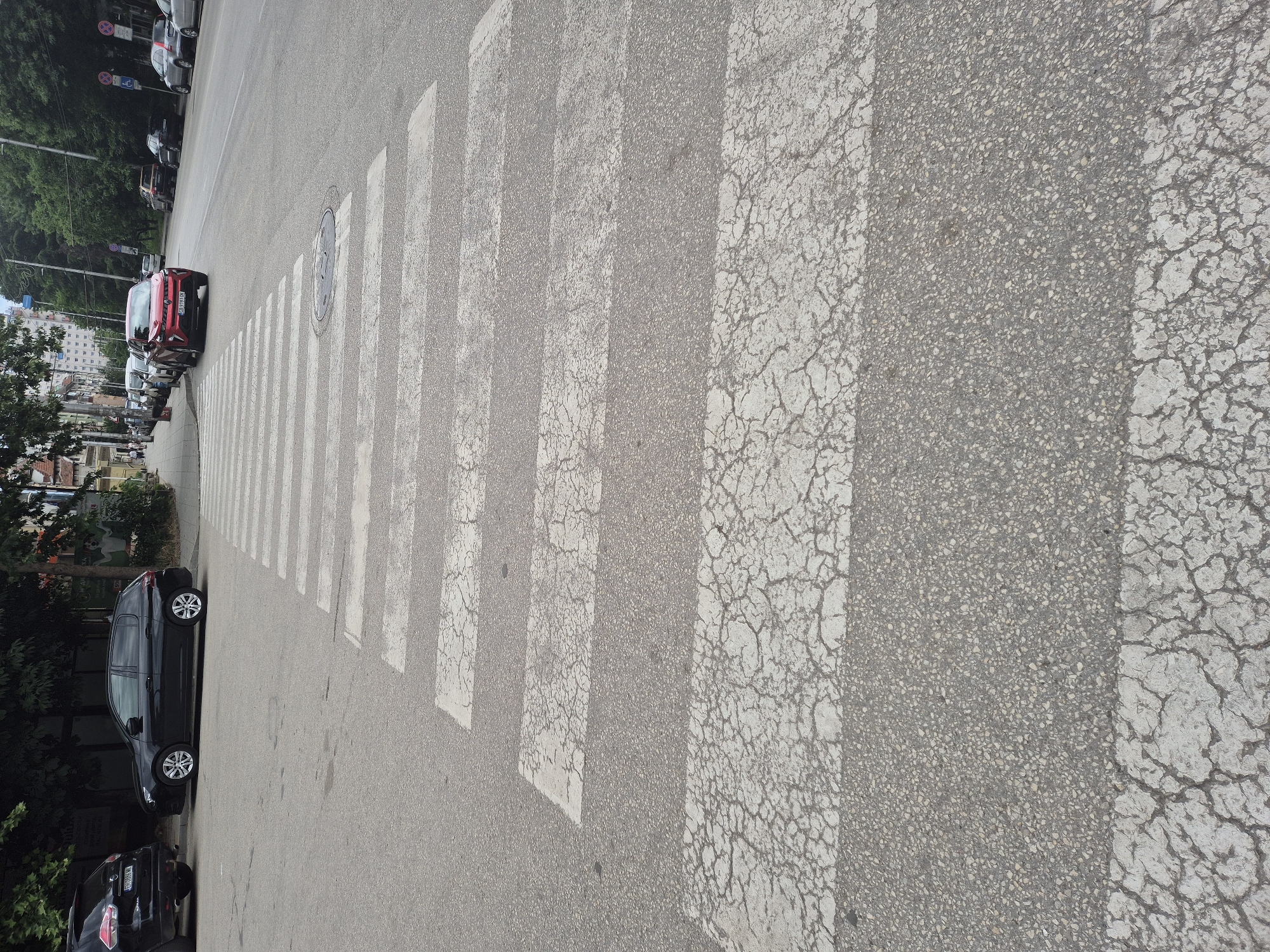 In an urban environment, a pedestrian crossing is prominently featured in the foreground. The crossing, composed of alternating white stripes and asphalt, shows signs of wear with cracks appearing in the white paint. Beyond the crossing, several cars are parked along the side of a street, with more vehicles visible in the distance. Trees line the street, and buildings are present in the background, suggesting a city setting. The sky is overcast, and the overall lighting indicates daytime.