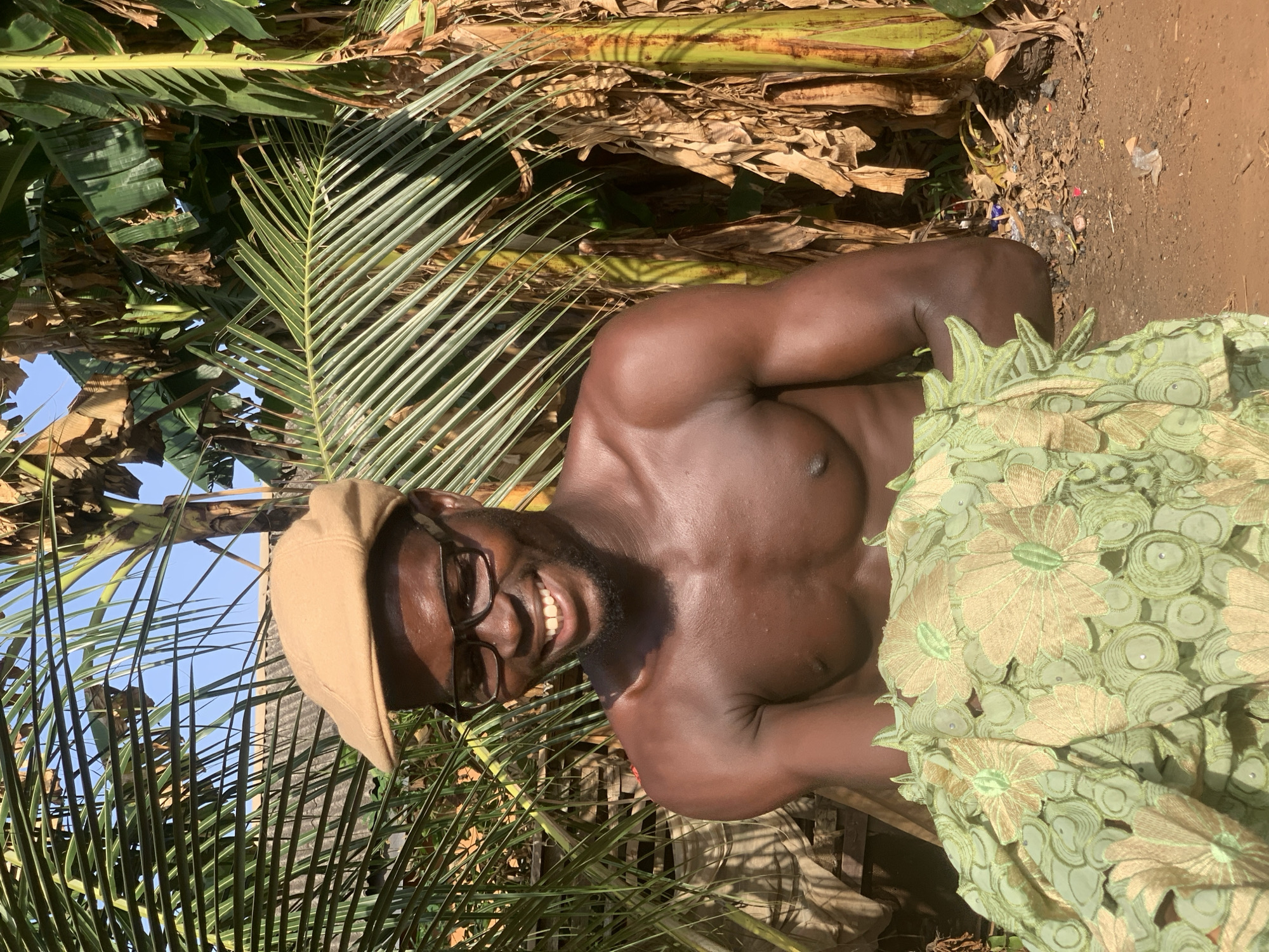The image shows a shirtless man with dark skin, wearing a tan cap and glasses, smiling towards the camera. He is partially covered by a piece of light green fabric with a floral pattern. The man is surrounded by palm fronds. The background also features dried banana leaves. A patch of bare earth with some litter is visible. The location is indicated to be Ikorodu, Nigeria, implying an outdoor setting with tropical vegetation.