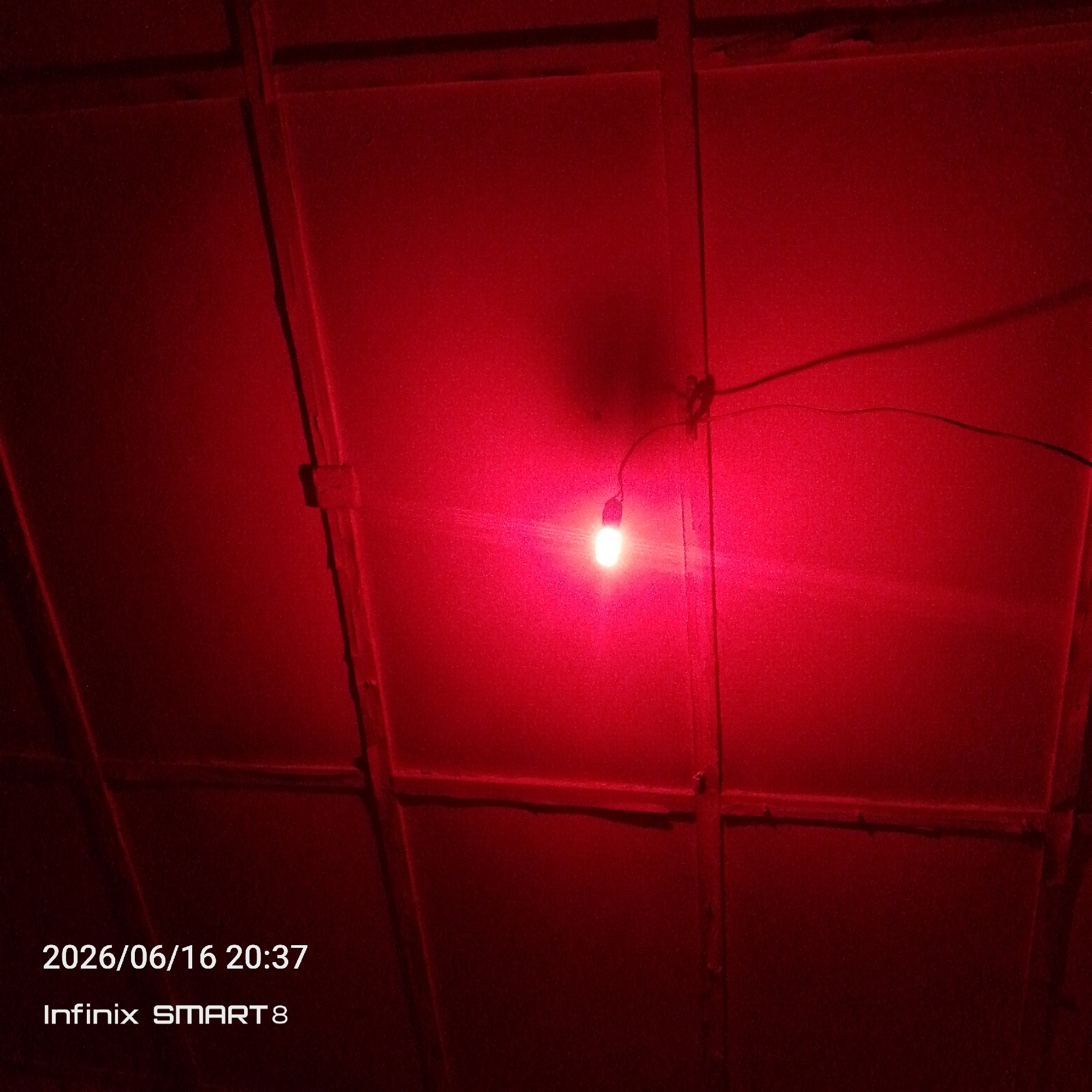 The image shows a red light bulb hanging from a wire in what appears to be a dimly lit, indoor space. The ceiling is constructed with wooden beams and panels, typical of some structures in Nigeria. A timestamp in the lower left corner indicates the photo was taken on 2026/06/16 at 20:37, and identifies the device used as an "Infinix SMART 8". The overall ambiance is dark and the red light casts a strong, uniform hue across the scene, obscuring finer details of the environment. There are no people visible in the image.