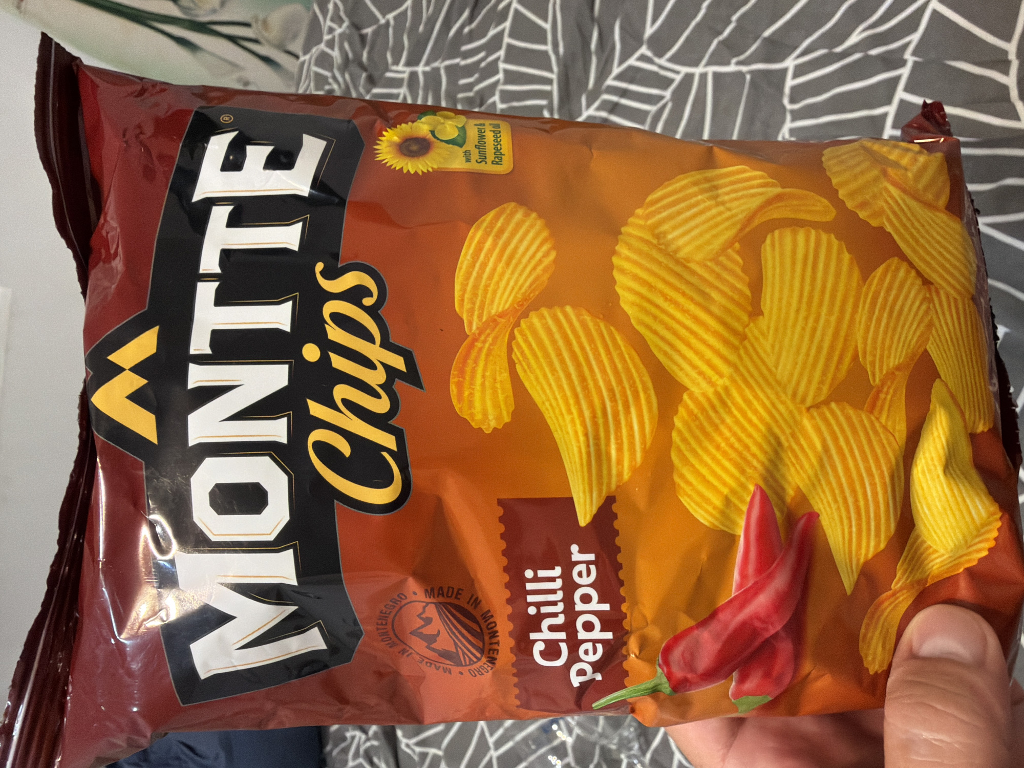 A close-up, indoor shot shows a person's hand holding a bag of "MONTE Chips" with a "Chilli Pepper" flavor. The bag is predominantly red and orange, with yellow ridged potato chips depicted as part of the design, along with two red chili peppers. The bag also features a circular logo that says "MADE IN MONTENEGRO" with a mountain and field illustration. A small yellow sunflower and leaves are shown with the text "with Sunflower & Rapeseed oil". The background is a grey and white patterned fabric. The item is a packaged snack product.