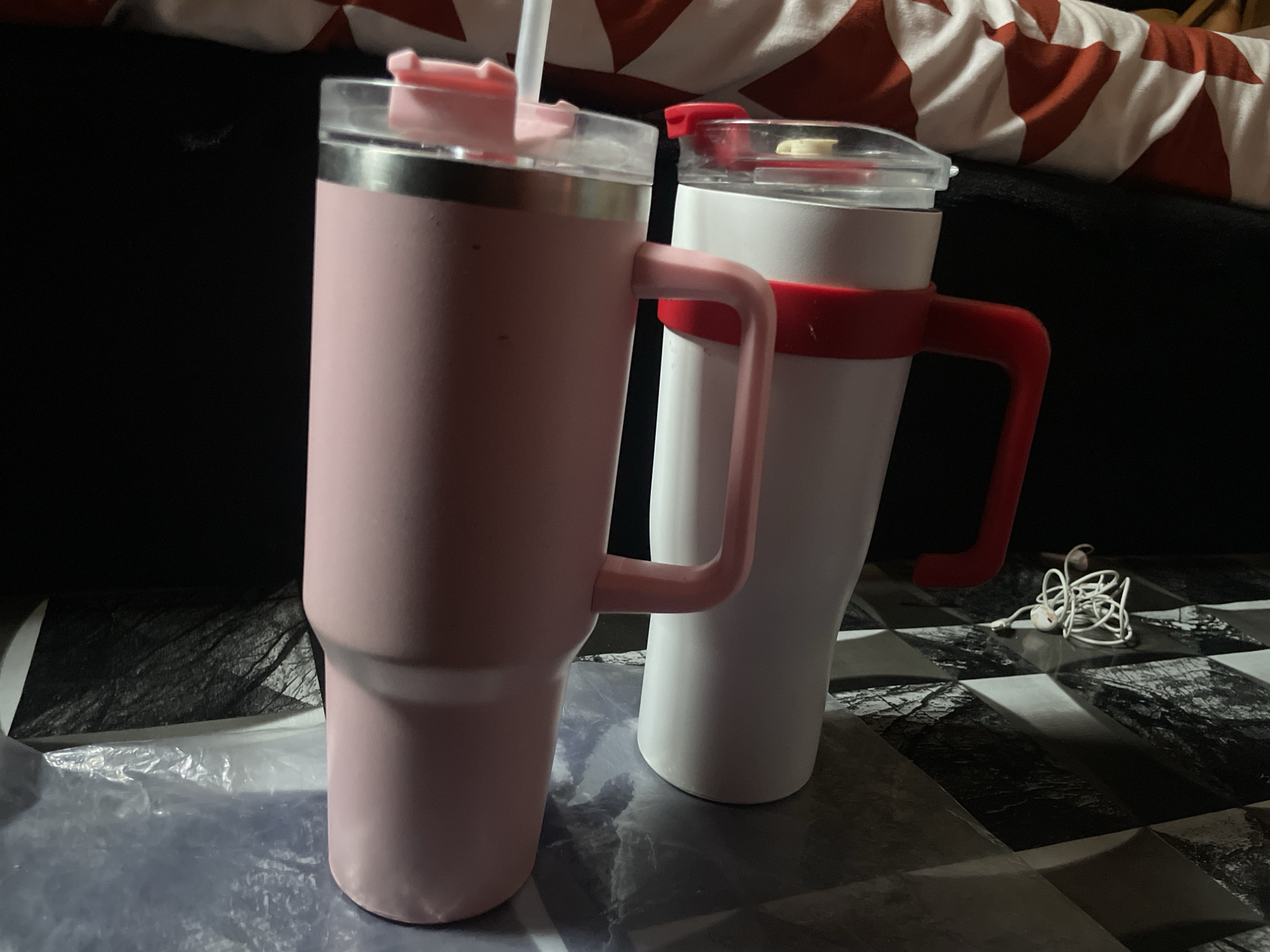 Two large, insulated tumblers with handles and lids are placed on a checkered surface. The tumbler on the left is pink and has a white straw. The tumbler on the right is white with a red handle and lid accent. A coiled white earphone cord is on the surface to the right of the white tumbler. The background is dark and out of focus, with a patterned fabric visible at the top. There is no indication of the location being in Jalingo, Nigeria.