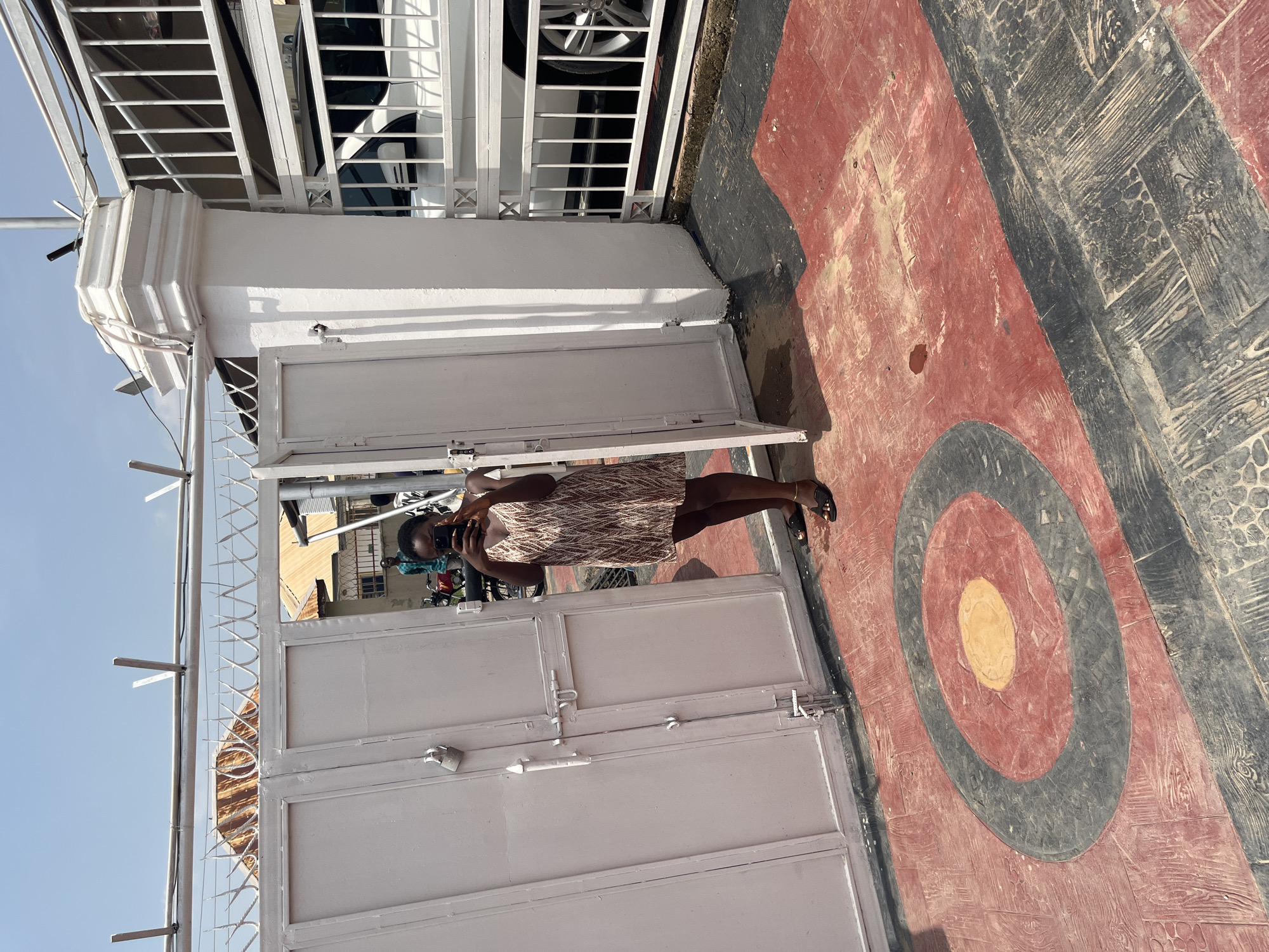 A young Black woman stands by a white metal gate, holding a phone and taking a selfie. She is wearing a short, patterned dress and sandals. The gate has a lock and appears to be the entrance to a property. Behind the gate, a white car is partially visible. The ground in front of the gate is decorated with a large, circular target-like design in red, black, and yellow. The scene appears to be outdoors, with a clear blue sky and bright sunlight, suggesting a sunny day. The overall setting suggests a residential or commercial property entrance in an urban environment, with a slightly weathered and textured appearance to the ground and gate. The woman's posture and focus on her phone indicate she is actively engaged in taking the photograph.