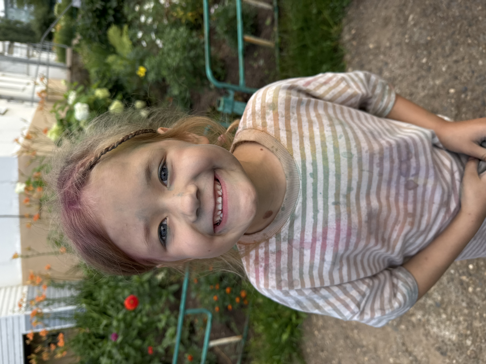 A young girl with blue eyes and light brown hair, some of which is dyed pink, is smiling at the camera. Her face is smudged with dirt or paint. She is wearing a striped t-shirt with pastel colors. The background is a garden with flowers and a white building. The girl appears to be in an outdoor setting, possibly a backyard or park, in Chaikovsky, Russia.