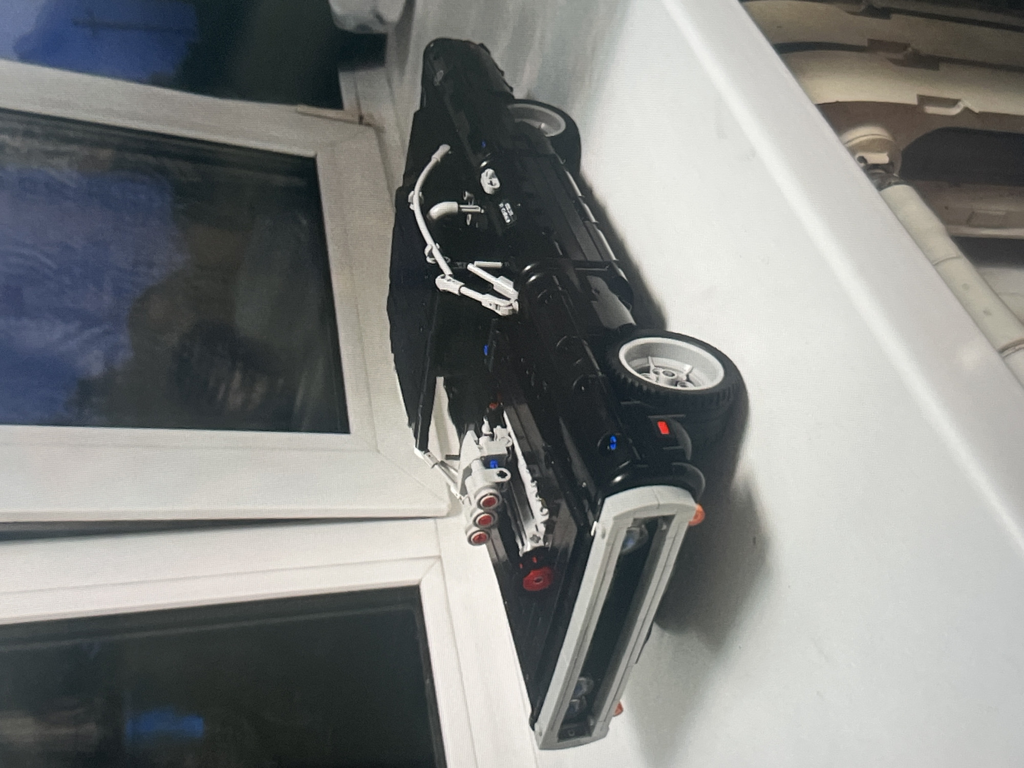 This is a close-up, eye-level, slightly angled photograph of a black LEGO replica of a 1970 Dodge Charger R/T, likely the one from the Fast & Furious franchise. The model is placed on a white, flat surface, which appears to be a windowsill or ledge. Behind the model, there are white window frames and dark tinted windows, through which a dim, blueish-grey sky can be seen, suggesting it is either dusk or a cloudy day. The model is detailed, featuring a visible engine supercharger with red-tipped exhausts on the hood, a roll cage, and silver rims on the wheels. The lighting is soft, casting a gentle shadow to the right of the car. There is no indication of the city of Ob, Russia, or any urban environment; the scene appears to be indoors. There are no people present in the image.