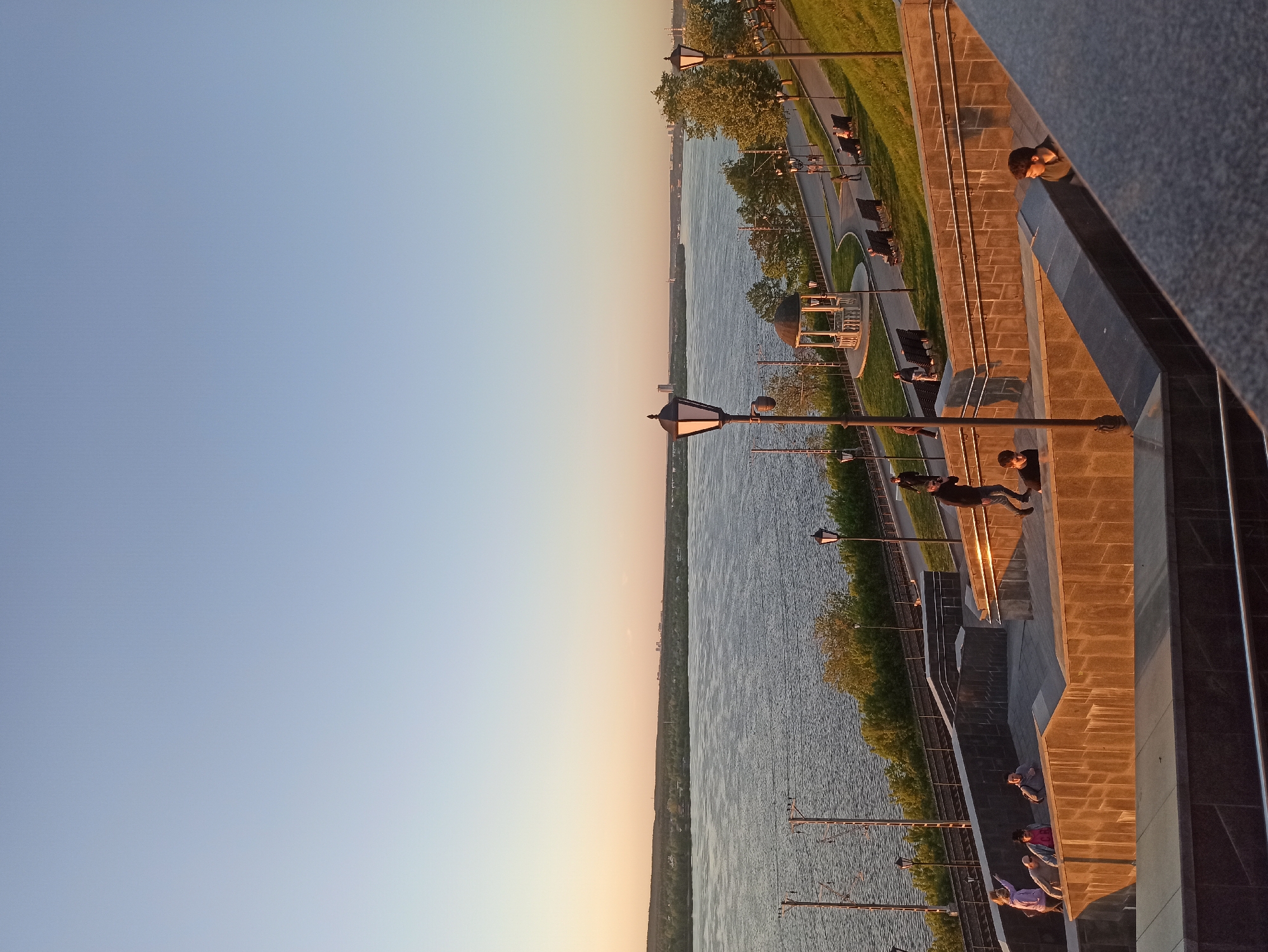 The image shows a waterfront scene in Unknown, Unknown. The sky transitions from blue to orange hues, indicating either sunrise or sunset. A body of water runs vertically through the center of the image. On the right is a paved area with trees, lampposts, benches, and a gazebo. People are walking along the walkway and sitting on benches. The right edge of the image shows a stone structure with steps. People are standing on the steps.