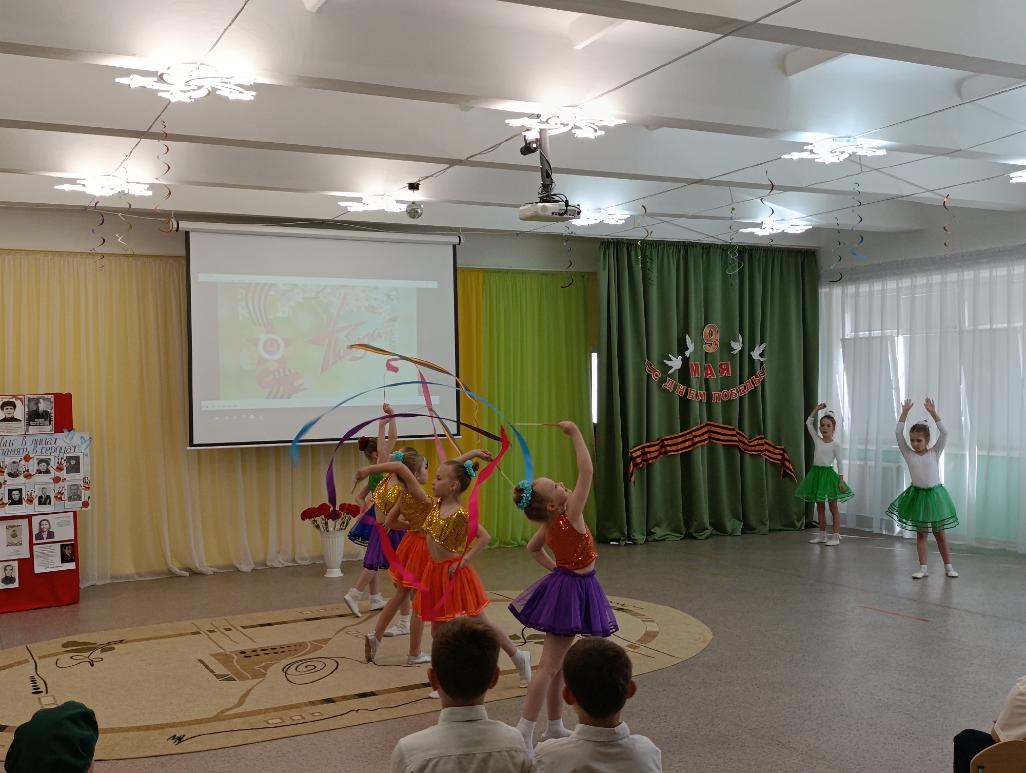 The image depicts an indoor event in what appears to be an auditorium or large room, possibly in Chaikovsky, Russia. Several young girls are performing a dance routine. At the center of the image are five girls with ribbons. They wear costumes consisting of sequined tops and colorful skirts. To the right, two other girls in white long-sleeved tops and green skirts are positioned near windows. In the foreground, two boys are seated, facing the performance, and seem to be watching the performance. Decorations, including snowflake-shaped light fixtures and colorful streamers, adorn the ceiling. A projector is suspended from the ceiling. A projection screen displays an image. To the left, a display board showcases photographs and text, likely related to an event or commemoration. Green curtains are on the right side, on which the words "9 May" are written in Russian, along with other text and symbols associated with Victory Day celebrations. The room has a beige rug with a geometric pattern.