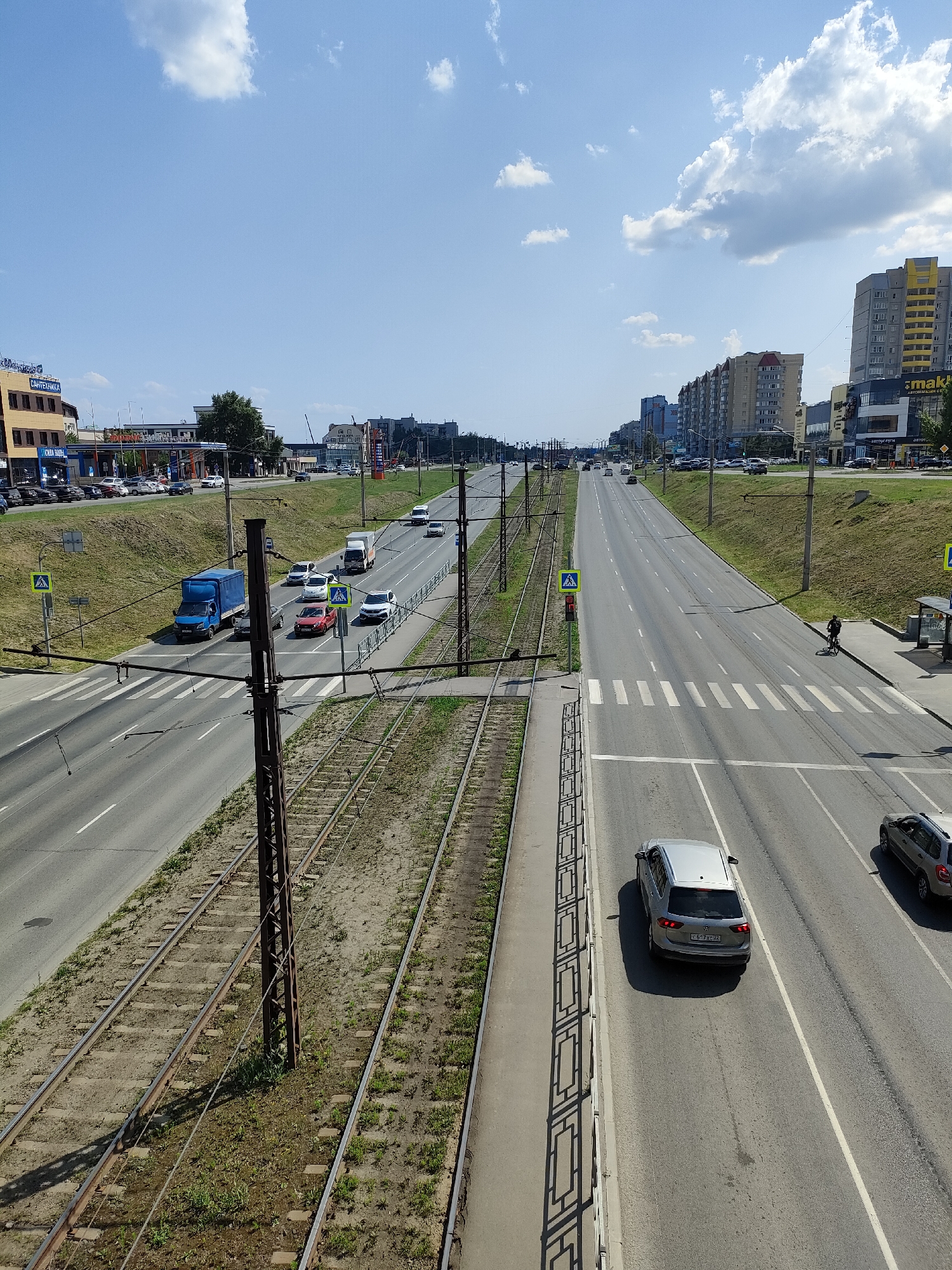 The image depicts a wide, multi-lane road with tram tracks running alongside it, under a bright, partly cloudy sky. The scene appears to be in an urban or suburban area, possibly Barnaul, Russia, as suggested by the overall setting and architecture.
On the road, various vehicles are in motion. A blue truck is seen on the left side, near a pedestrian crossing. Several cars, including a silver SUV and a darker SUV, are driving in the right lanes. A person on a bicycle is visible on the sidewalk on the right side of the road, near a bus stop. The tram tracks are visible in the foreground, with overhead power lines supported by metal poles.
Buildings line the sides of the road. On the left, there are commercial buildings with signage, including what appears to be "Santehnika" and "Aqua Mira." On the right, taller apartment buildings are visible, some with yellow accents. One building has a large sign that reads "mak."
The time of day appears to be daytime, likely midday, given the bright sunlight and the shadows cast by the vehicles and poles. The weather is fair with scattered clouds. The overall atmosphere is that of a typical day with traffic and urban activity. The mood is neutral, focused on documenting the scene.