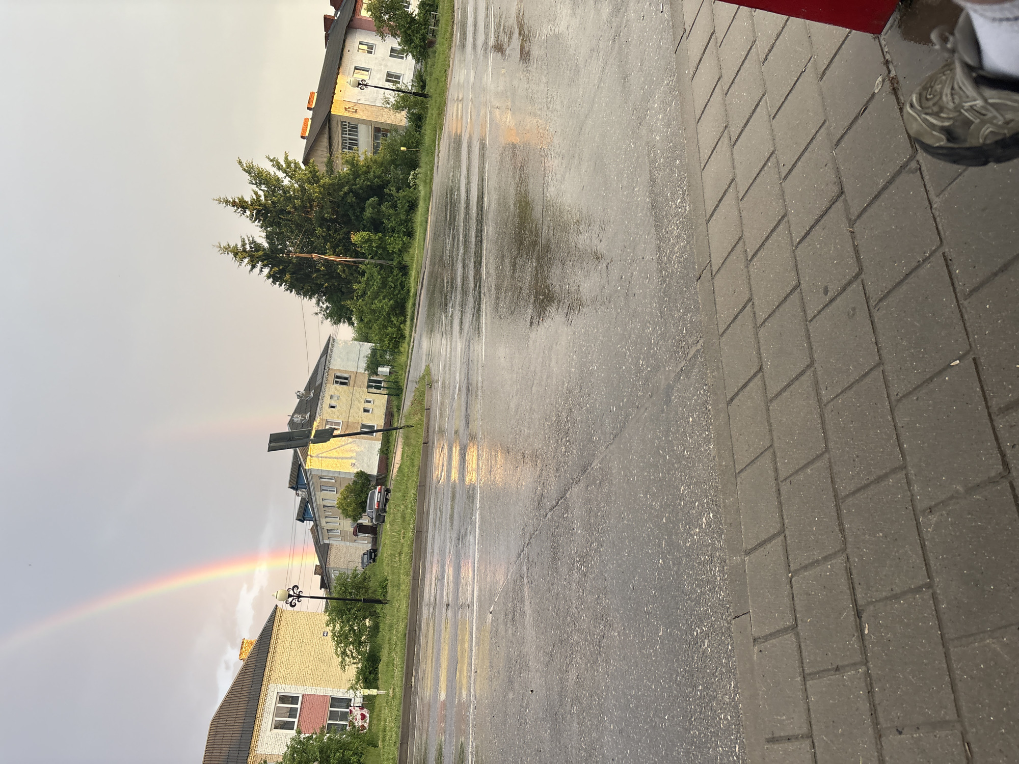 This image depicts a wet street scene in the city of Unknown, Unknown, under a cloudy sky with a double rainbow. The foreground shows a paved sidewalk made of rectangular concrete slabs, with a person's foot in a gray/green sneaker and white sock visible in the bottom right corner. Adjacent to the sidewalk is a dark asphalt road, thoroughly wet and reflecting warm light (yellow, orange, pink) from the sky and the visible double rainbow. The primary rainbow is vibrant and distinct, while the secondary rainbow is fainter.
To the left of the road, a grassy verge separates the street from residential buildings. A two-story building with cream-colored walls, a dark sloped roof, and a distinct red/pink section on its facade is visible. A decorative black street lamp post stands in the grass near this building. Further down the road, a dark sedan is partially visible behind the lamp post.
On the right side of the road, more buildings are present, ranging from two to three stories, displaying varied architecture including some with white walls and dark roofs, and others with cream or yellow facades. Tall, dark green coniferous trees are centrally located, partially obscuring some structures. A large, dark rectangular sign or board is visible on the grassy verge to the right. Another dark sedan is parked further along the road on the right. The overall impression is of an urban or semi-urban environment shortly after rainfall, with the sun breaking through to create a rainbow and warm reflections.