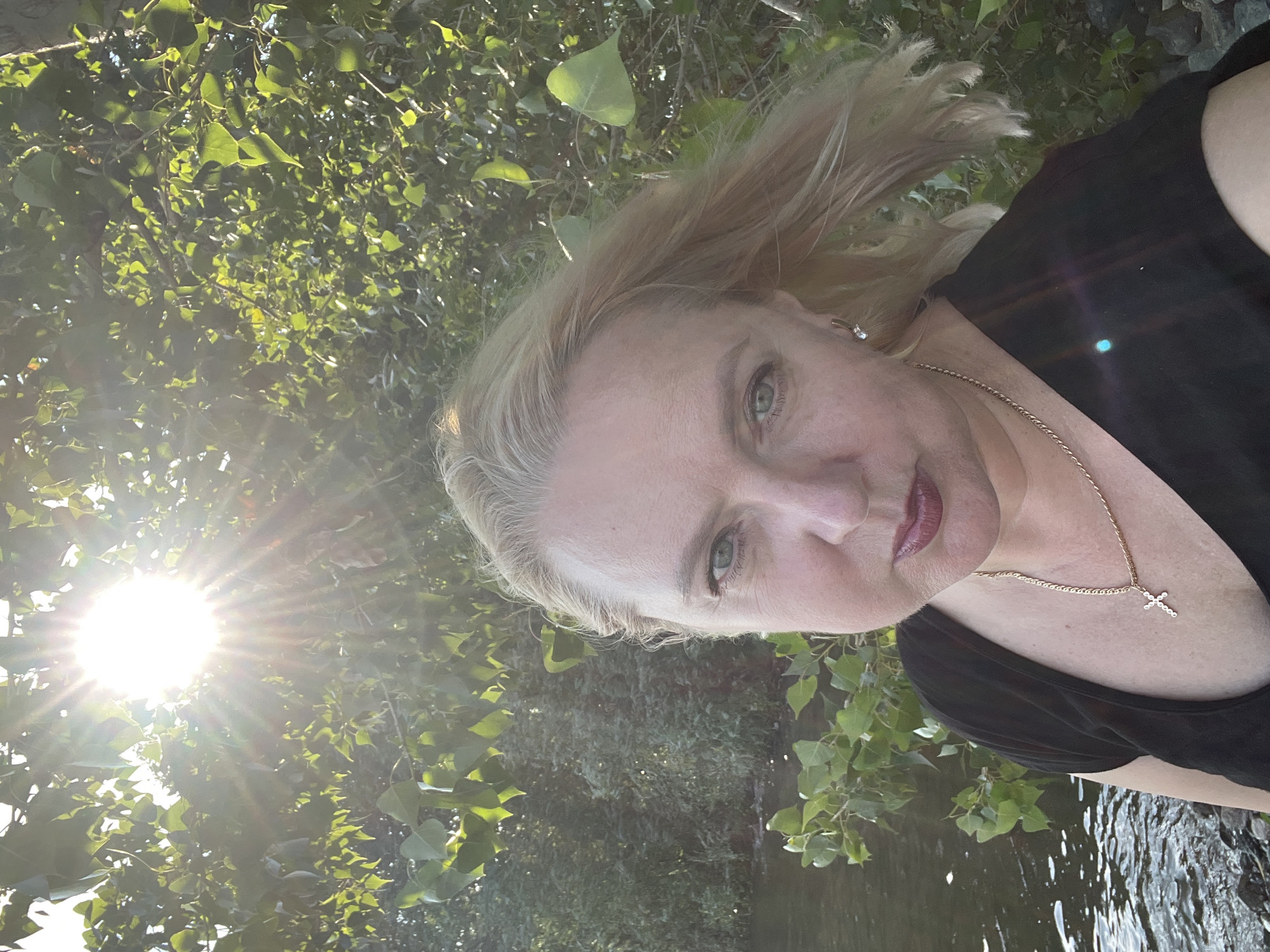 A woman with blonde hair and blue eyes is standing in front of a body of water and trees. She is wearing a black shirt and a gold necklace with a cross pendant. The sun is shining brightly through the trees, creating a lens flare effect. The image appears to have been taken outdoors in a natural setting. It is not possible to determine the specific location in Kazan, Russia, from the image alone.