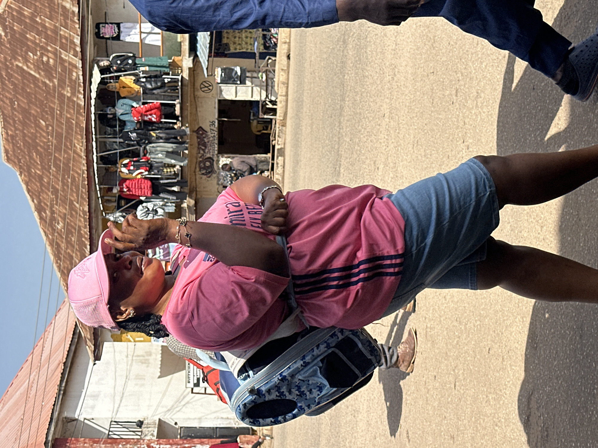 In Bukuru, Nigeria, a woman walks on a dusty street. She wears a pink t-shirt, a pink cap, and denim shorts, with a blue and white patterned bag slung over her shoulder. Her left hand is raised to her face, and she wears a watch and a bracelet. A man in a blue shirt walks past her on the right. In the background, a market stall displays various jackets and clothing items on mannequins. Buildings with corrugated iron roofs line the street. The scene is bathed in bright sunlight, casting shadows on the ground.