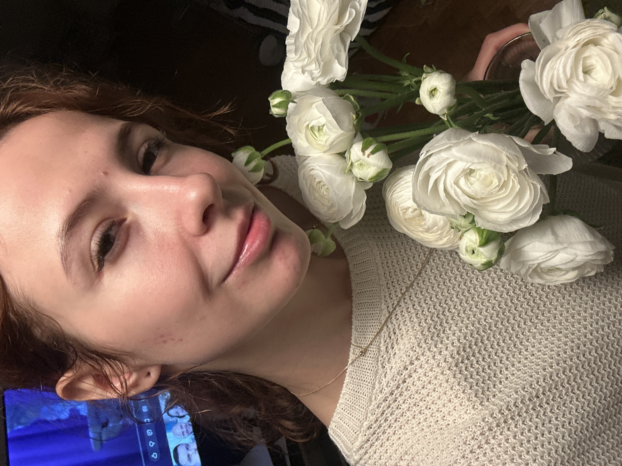 A young woman with reddish-brown hair is lying down, wearing a light beige knitted sweater and a delicate gold necklace. She is partially illuminated, with the light casting shadows on her face. She is holding a bouquet of white ranunculus flowers close to her face and body. A television screen is visible in the lower left corner, displaying images of people. The location of the photo cannot be identified from the image provided as Novi Sad, Serbia.