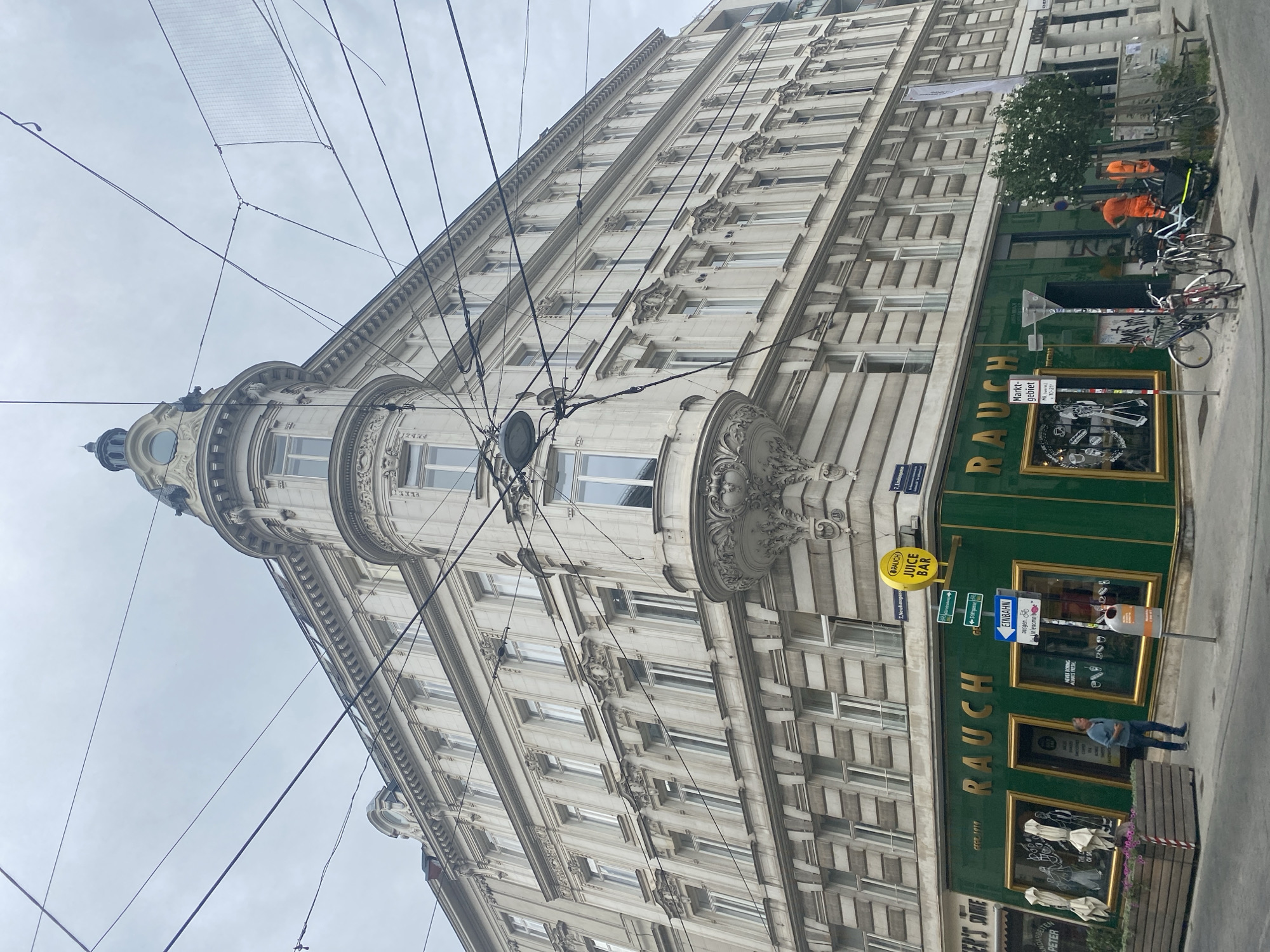 This image depicts a street-level view of a building with a green storefront at the corner of a street in Vienna, Austria. The building is an ornate, multi-story structure with cream-colored facades and many windows. The ground floor features a prominent business with the word "RAUCH" in large gold lettering across its green facade. The storefront appears to be a juice bar, as indicated by a yellow sign that reads "RAUCH JUICE BAR" and window displays showing graphics related to juices and healthy eating.
The street is paved, and there are overhead electrical wires and tram lines crisscrossing the sky, typical of Viennese urban infrastructure. Several street signs are visible, including one with an arrow pointing to "Rathaus" and "Neubaugasse," indicating a location in the Neubau district of Vienna. There are bicycles parked in front of the juice bar, and a few people are present. One man in a blue shirt stands on the sidewalk in front of the store, while two individuals in bright orange work attire are near the entrance on the right side of the frame, conversing next to a tree. Planters with flowers are visible on the left side of the storefront. The overall atmosphere is that of an active urban street scene in a European city.