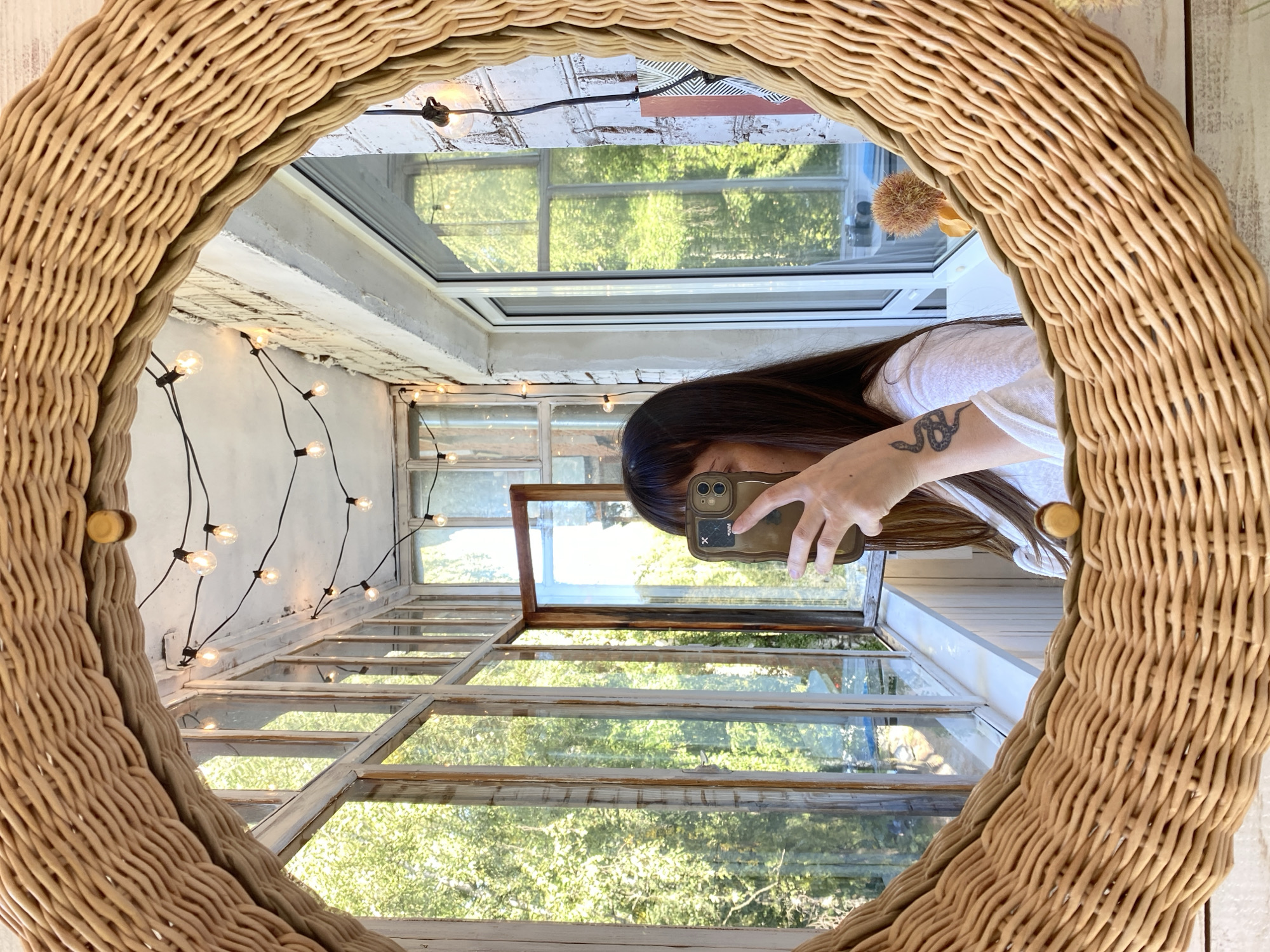 A person, partially obscured by a smartphone held to take a mirror selfie, is reflected in a wicker-framed oval mirror. The person has long dark hair and is wearing a white top. A snake tattoo is visible on their left arm. The mirror is positioned on a wooden surface, possibly a table, and reflects a sunlit enclosed balcony with large windows. Through the windows, lush green trees are visible, suggesting a location with abundant vegetation. String lights with exposed bulbs are draped across the ceiling of the balcony. The architectural style of the windows and the presence of a brick wall indicate an older building. The scene appears to be in Votkinsk, Russia, given the context.