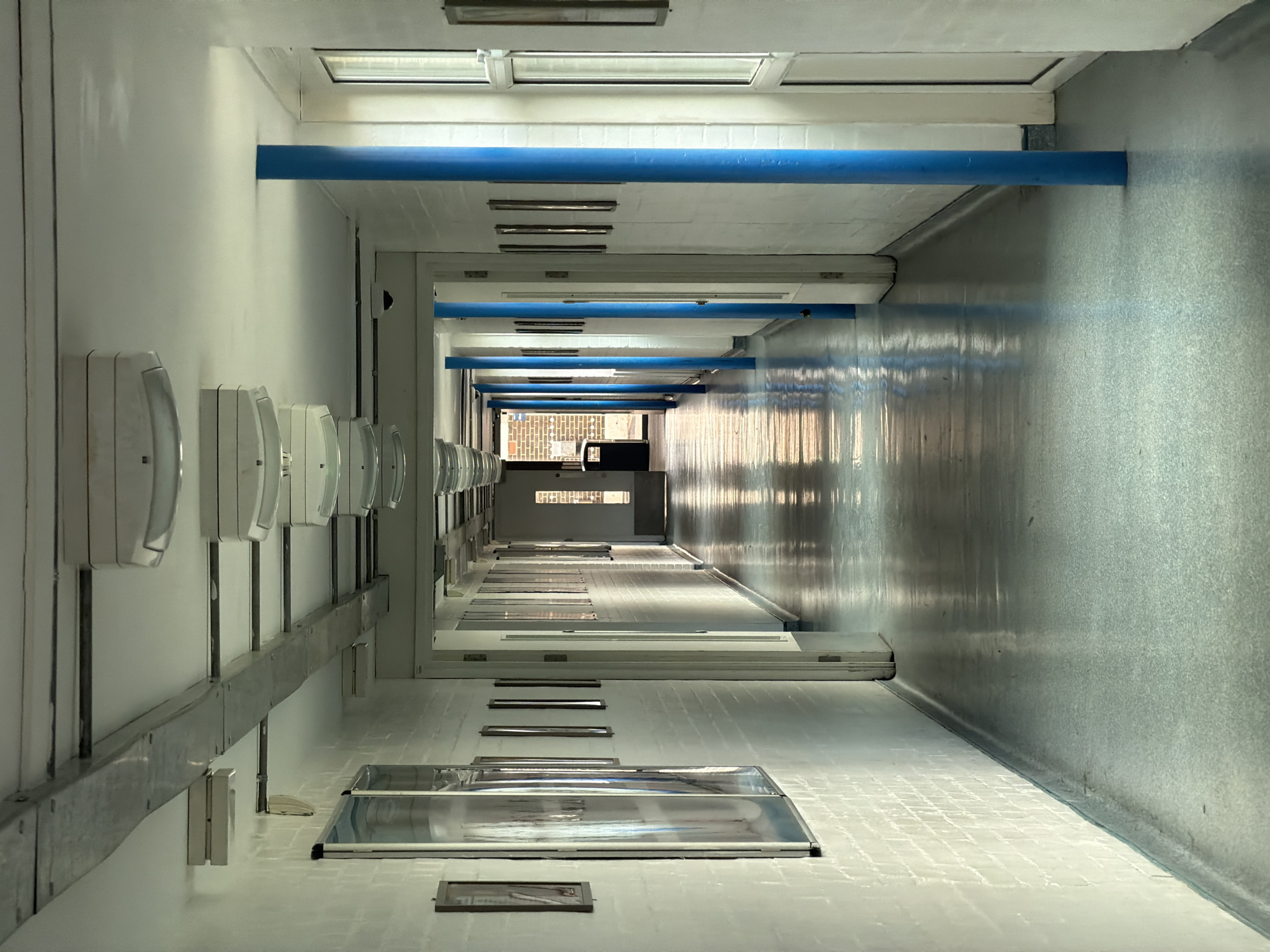 The image depicts a long, empty interior corridor within a building in London, United Kingdom. The corridor features white-painted brick walls. The left wall displays a series of framed posters or prints, some covered in clear protective sheeting, along with smaller framed pictures. The right wall presents a repeating sequence of white doors with adjacent glass panels or windows. A series of bright blue cylindrical pillars are positioned along the right side of the corridor, extending from the light grey-blue, highly reflective linoleum floor to the white ceiling. The ceiling is fitted with multiple elongated fluorescent light fixtures and a prominent grey metallic duct or cable trunking runs along the upper left. Natural sunlight streams from an open doorway at the far end of the corridor, illuminating the reflective floor and casting distinct shadows. A dark waste bin is visible near the doorway at the corridor's termination. No individuals or active subjects are present.