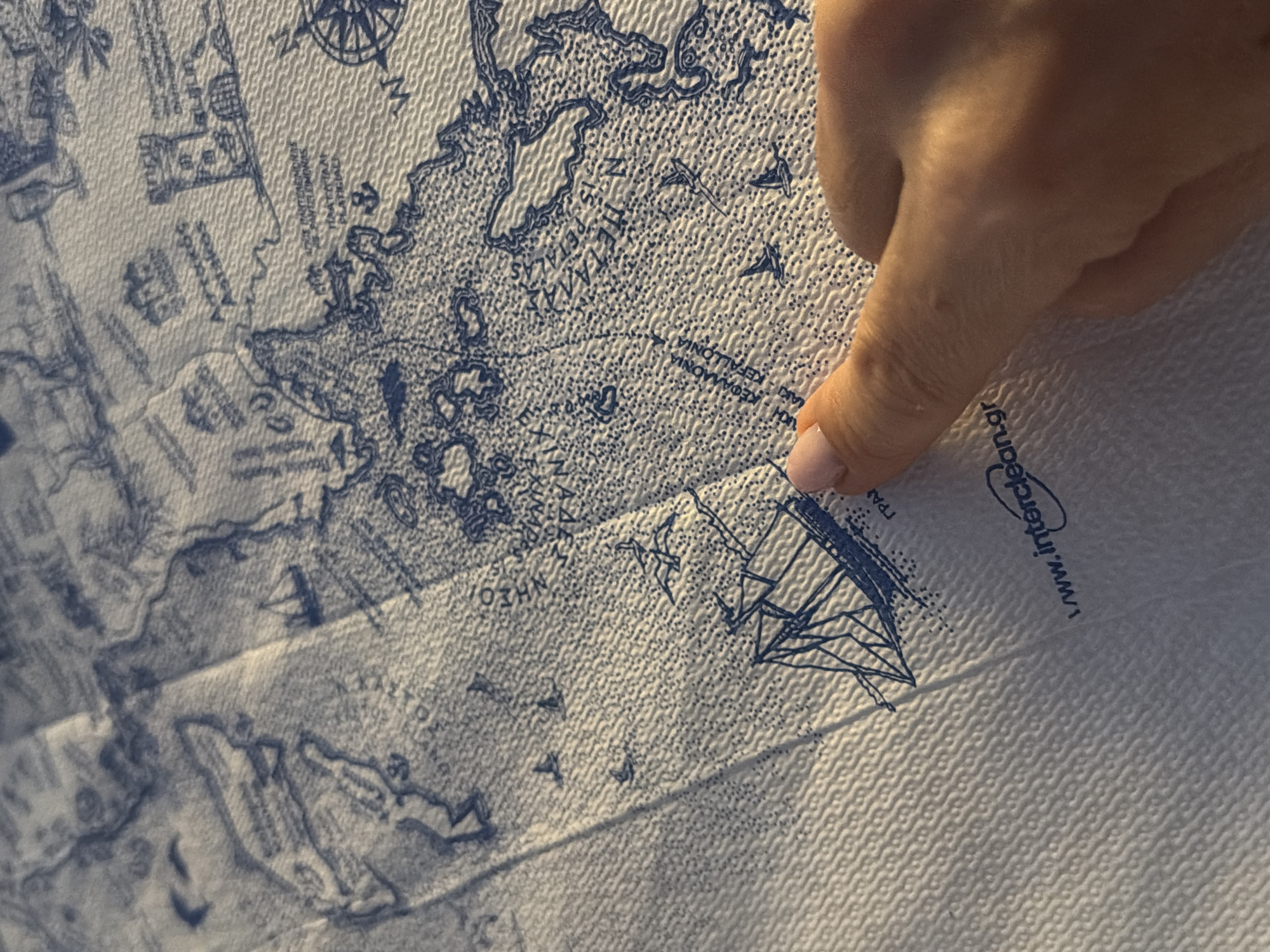 A close-up, overhead shot shows a person's thumb and index finger pointing at a blue and white map printed on a textured, white material. The map depicts a coastal area with islands, boats, and what appear to be buildings or ruins. Text in blue lettering is visible on the map. The bottom of the image features a portion of a logo with the text "www.interclean.gr". The lighting suggests a natural outdoor setting, possibly with sunlight casting shadows.
Based on the provided text on the map, "Municipal Unit of Alyzia, Greece" is a relevant location. Specifically, the map shows an area with islands and coastal features that could correspond to this region. The text on the map, though partially obscured, appears to include place names or geographical markers within Greece.