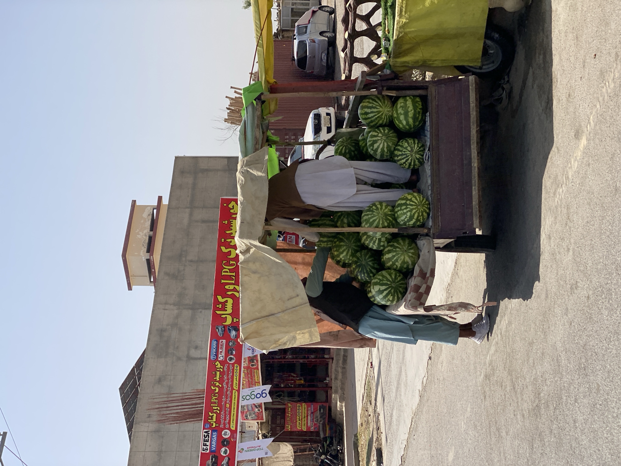 The image shows a street scene in Kandahar, Afghanistan. A cart filled with watermelons is parked on the side of the road, under a makeshift canopy. Two men are attending to the cart. One man, wearing a light blue shalwar kameez and white sneakers, is bent over, appearing to be arranging or selling the watermelons. The other man, wearing a light brown vest over a white shalwar kameez, is standing behind the cart. In the background, there is a building with a large red and yellow sign displaying text in what appears to be Pashto or Dari, advertising LPG and other services. Several vehicles are visible in the distance, including a white SUV and a silver car. The sky is clear and blue, suggesting a sunny day. The street surface is paved asphalt.