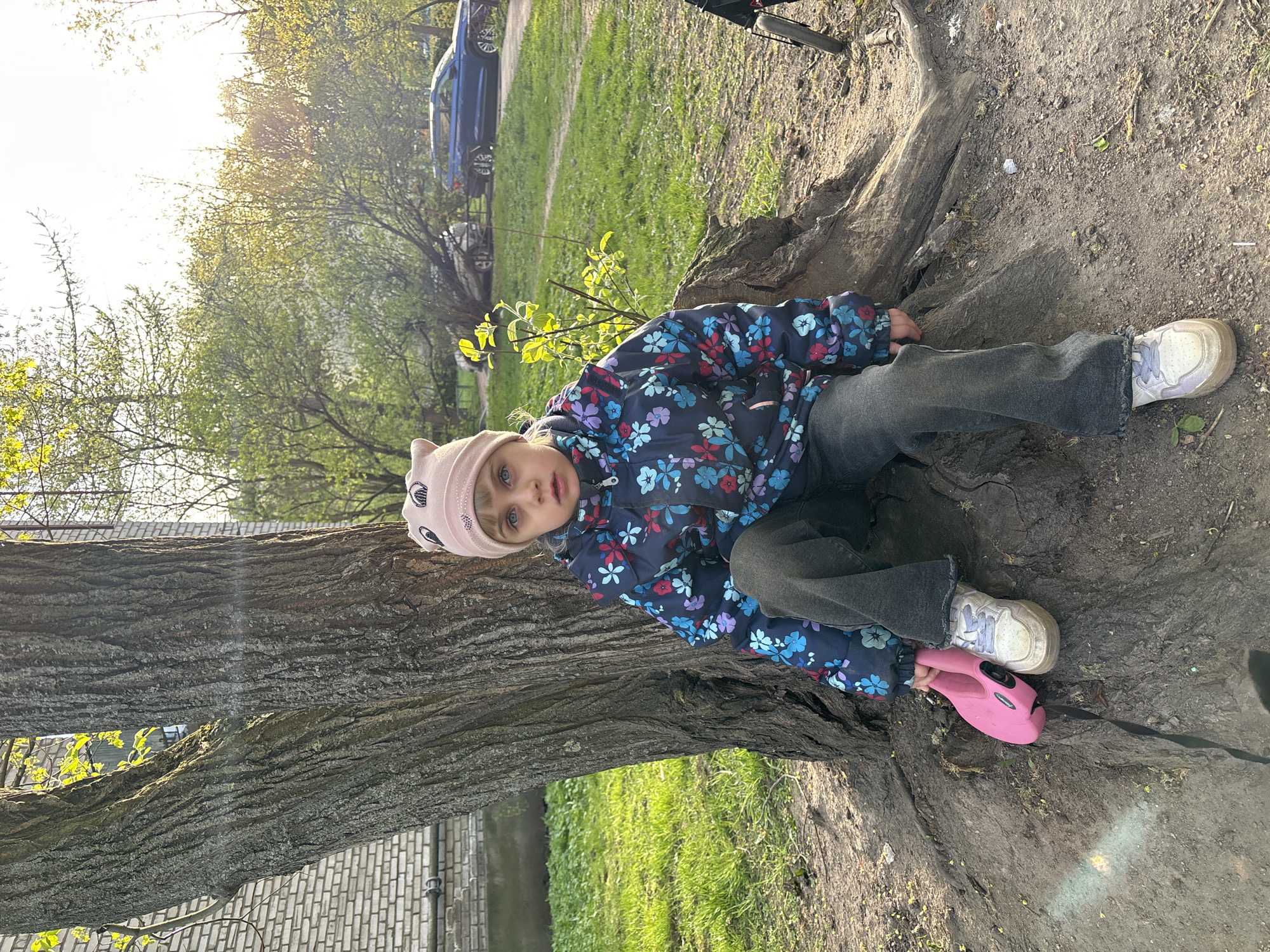 A young girl lies on the ground near the base of a large tree. She wears a floral-patterned jacket, gray pants, a pink hat, and white sneakers with a pink object next to her foot. The tree has a thick trunk and exposed roots. Behind the tree, there is green grass, other trees, and a glimpse of a blue car. A brick building is also partially visible. The ground is a mixture of dirt and tree roots. The lighting suggests it is daytime. The location may be the city of Saint Petersburg, Russia.