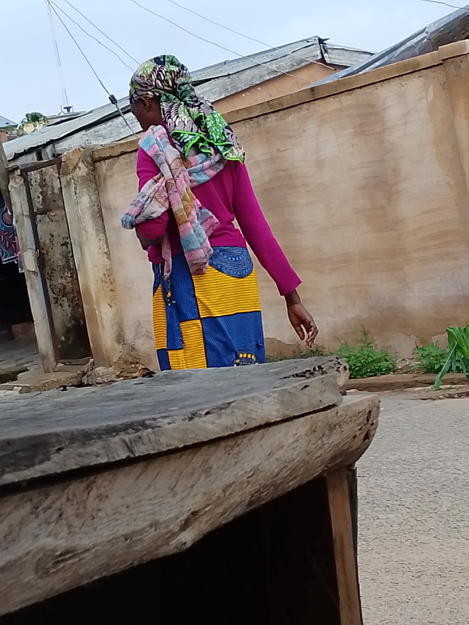 In a residential area of Jos, Nigeria, an individual with a patterned headscarf, pink long-sleeved shirt, and a patchwork blue and yellow skirt walks in front of a beige concrete building. A weathered, wooden structure occupies the foreground, partially obscuring the view. Vegetation grows near the wall and street.