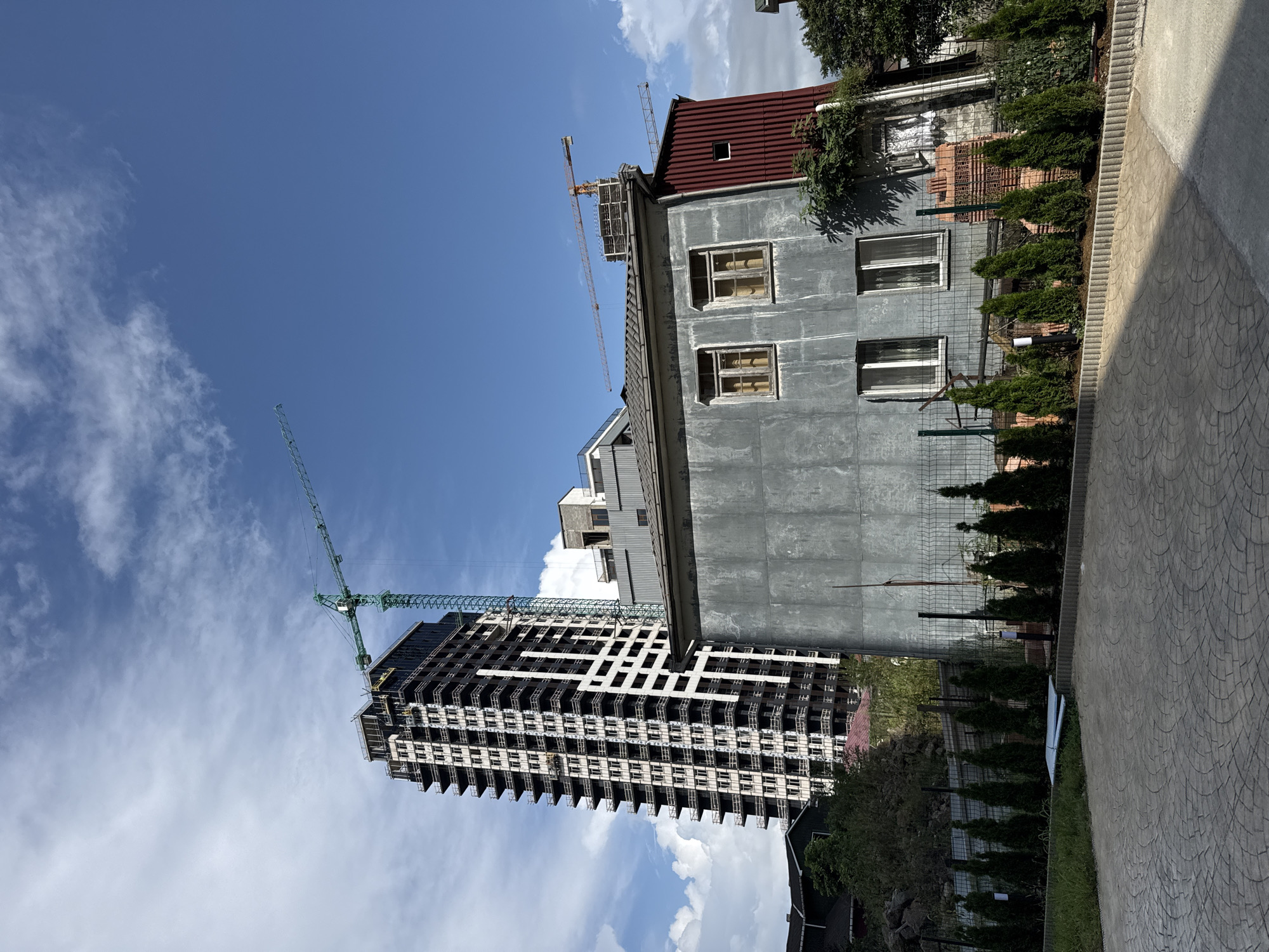 This is a photograph taken on a sunny day in Batumi, Georgia, featuring a stark contrast between old and new architecture. In the foreground, a paved area with a circular pattern leads towards a low fence lined with conical evergreen trees. Behind the fence is an older, gray-sided building with a red corrugated metal roof. This building has several windows on its upper floor.
Dominating the background is a modern, multi-story building under construction, characterized by its dark facade and numerous balconies with white railings. A tall green construction crane is positioned next to it, its boom extending over the building. Further in the background, another, smaller construction crane is visible on top of a more finished section of a building. The sky is a vibrant blue with scattered white clouds, suggesting fair weather. The scene captures a moment of urban development, where older structures coexist with new high-rises.