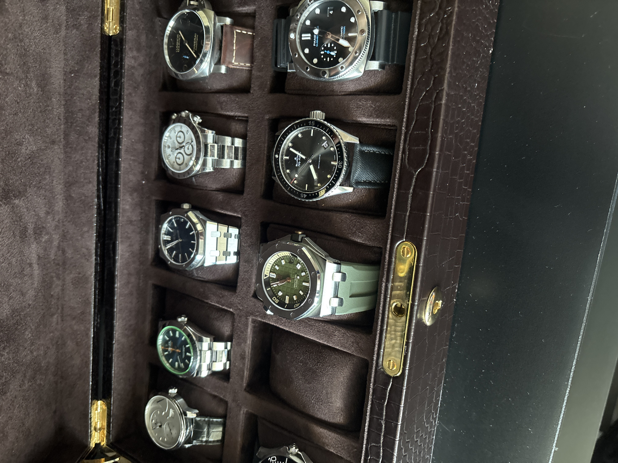 The image shows a dark brown, textured watch box with a gold clasp. The box contains ten watches arranged in two rows of five. The watches vary in design, with some having metal bands and others leather or rubber straps. The top row of watches, from left to right, includes a silver-faced watch with a black band, a watch with a green bezel and metal band, a watch with a black face and metal band, a silver watch with a white face and three subdials, and a watch with a black face and brown leather band. The bottom row of watches, from left to right, includes a watch with a black face and metal band, a watch with a green face and green rubber strap, a watch with a black face and black strap, and a watch with a silver face and black rubber strap. The watches are positioned in individual compartments within the box. The background is dark, suggesting an indoor setting. There is no discernible location information for Dubai, United Arab Emirates within the image.