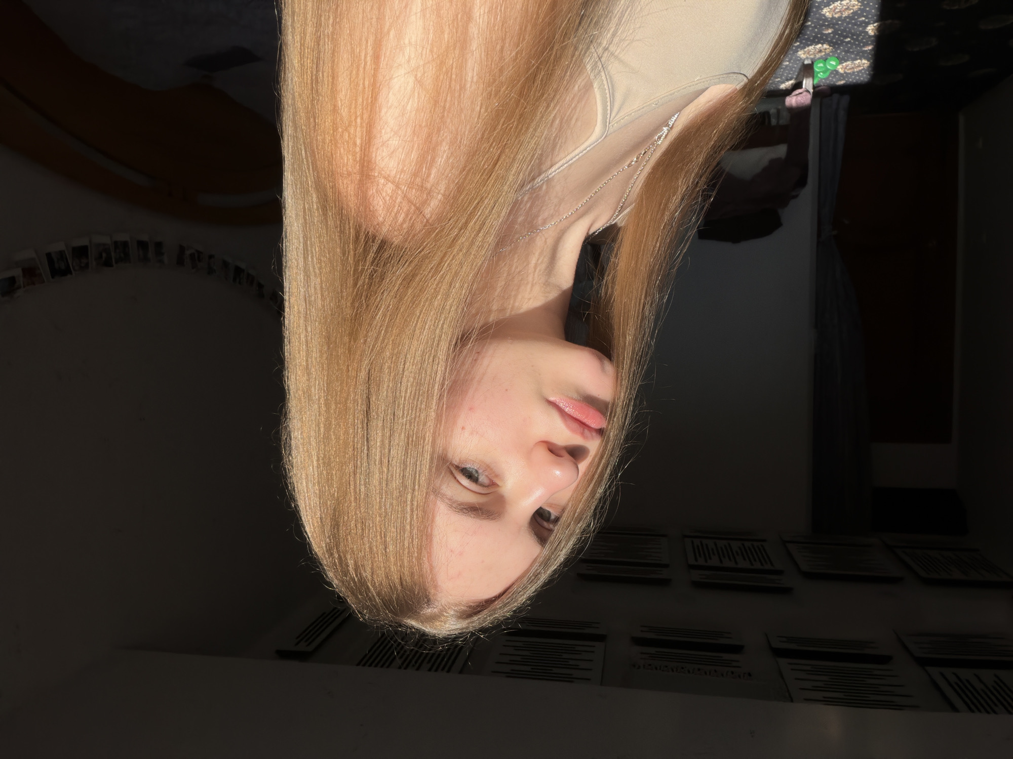 A young woman with long, straight blonde hair is shown from the chest up, posed in a sunlit room. She is looking towards the camera with a neutral expression and has light-colored eyes and clear skin. She is wearing a light-colored tank top and a silver necklace. Sunlight is casting shadows across the room, illuminating her face and hair. The background is dimly lit, with a bed visible on the right side of the frame and what appear to be acoustic panels on the ceiling. There are also small photos strung along the wall above the bed. The floor is carpeted with a patterned design. The scene suggests an indoor residential setting, possibly a bedroom, in Dnipro, Ukraine, given the prompt's location specification.