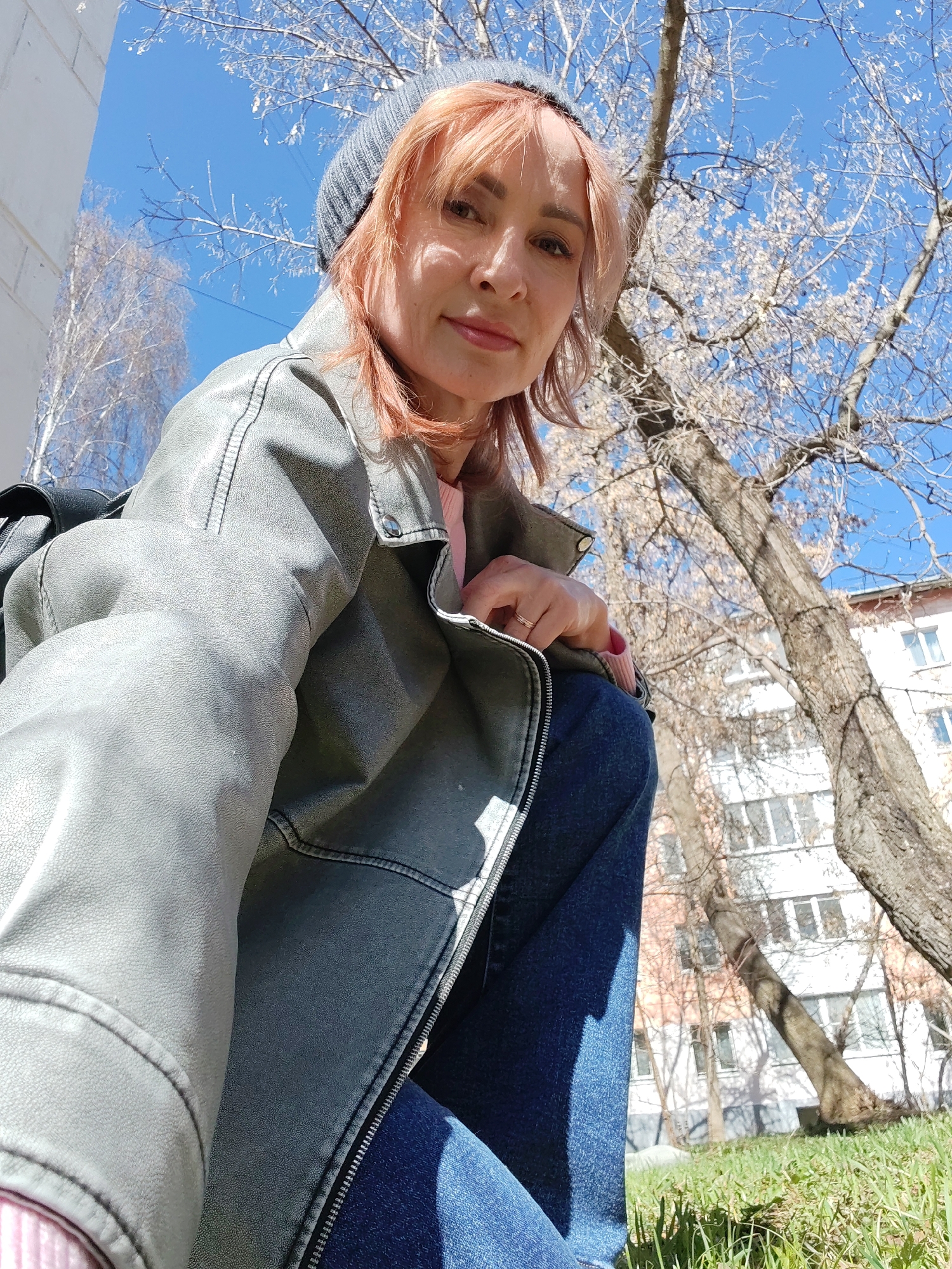 A woman wearing a gray beanie and a gray leather jacket is sitting outdoors on a patch of green grass. She is also wearing dark blue jeans and a pink sweater. She has fair skin and pinkish-red hair. In the background are trees with bare branches and a multi-story building with numerous windows. The sky is blue and clear. Based on the architecture and general appearance, it is likely a residential area in the city of Chaikovsky, Russia.