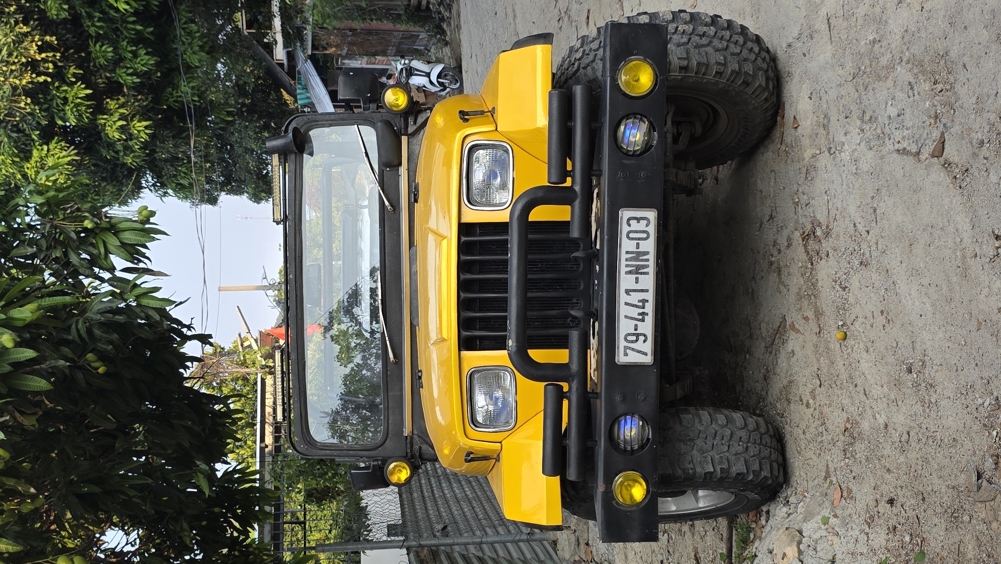 The image shows a yellow off-road vehicle parked on a rough, sandy surface. The front of the vehicle is prominent, featuring a black grill guard, large tires, and auxiliary yellow lights. The license plate is visible, bearing the number "79-441-NN-03". The background includes trees and vegetation, suggesting the location may be a natural or semi-rural environment. The area may be near the Bac Natural Heritage Area Trang, Vietnam, based on the landscape and vehicle type.