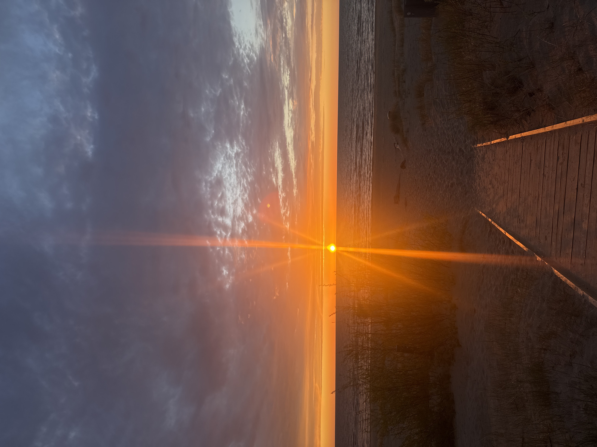 The image captures a sunset at the beach in Lilaste, Latvia. The sun is positioned at the horizon line, creating a bright orange glare that extends horizontally across the image. The sky is filled with a mix of dark, cloudy formations to the left, transitioning to softer, orange hues towards the center. The sea is visible on the right side of the image, reflecting the orange glow of the sunset. The beach area is overgrown with vegetation, leading to a wooden boardwalk in the foreground. No people are directly visible in the image.