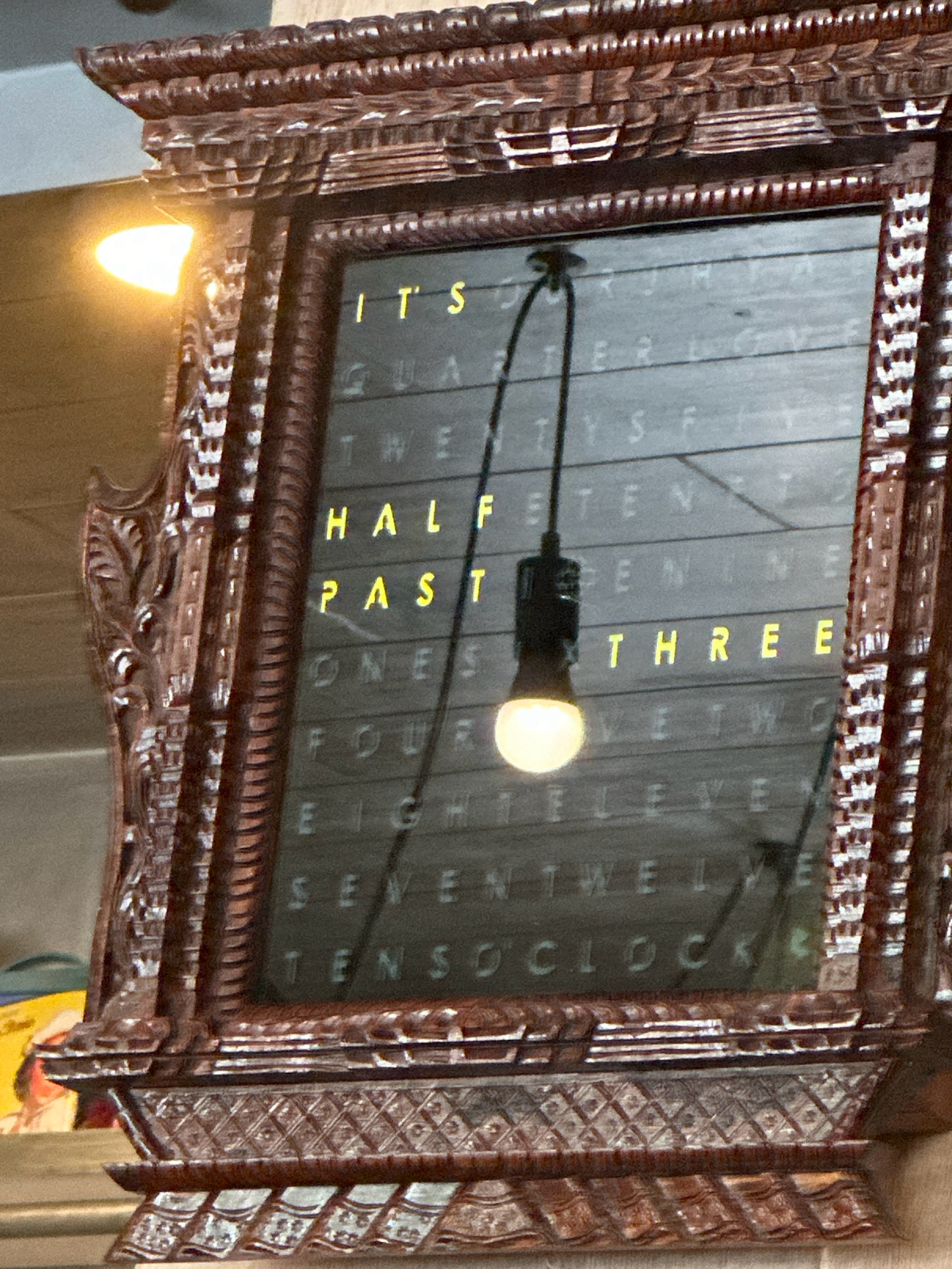The image shows a wooden-framed clock with a digital display that shows the time in words. The clock is hanging on a wall. The time displayed is "IT'S HALF PAST THREE". The clock face has a dark background with yellow letters that light up to show the words. There is a black cord with a lightbulb hanging from the top of the clock face. The clock is ornate and appears to be antique. The background of the image is blurry, but it appears to be an indoor setting. There are no people visible in the image. The location is not specified in the image or its metadata.