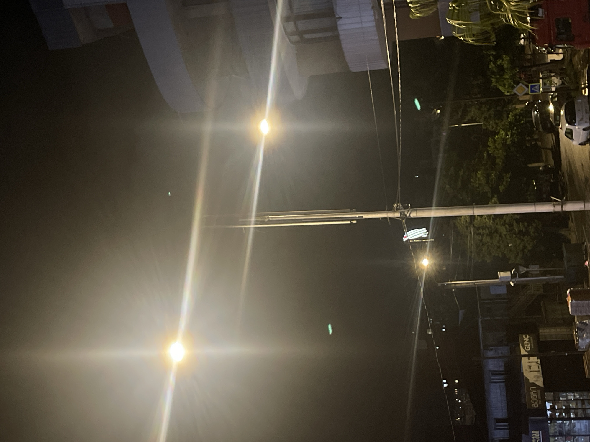 This image depicts a nighttime street scene in Batumi, Georgia, likely on a slightly overcast or foggy evening, indicated by the lens flares and diffused lighting. The prominent features are streetlights casting bright beams and creating starburst effects. In the background, a modern building with multiple floors and balconies is visible on the right, while a lower-rise building with visible windows and signage, including the text "GEN C", is present on the left. The street appears to be wet, suggesting recent rain. Several vehicles, including a white car and at least one dark SUV, are parked or driving on the street. There are also trees lining the street and what appear to be traffic signs, including a diamond-shaped yellow sign with a black symbol, and a white sign with a blue triangle and a black symbol. A red vehicle is partially visible on the far right. There are no discernible individuals in the image. The overall impression is a typical urban street at night, enhanced by the atmospheric lighting conditions.