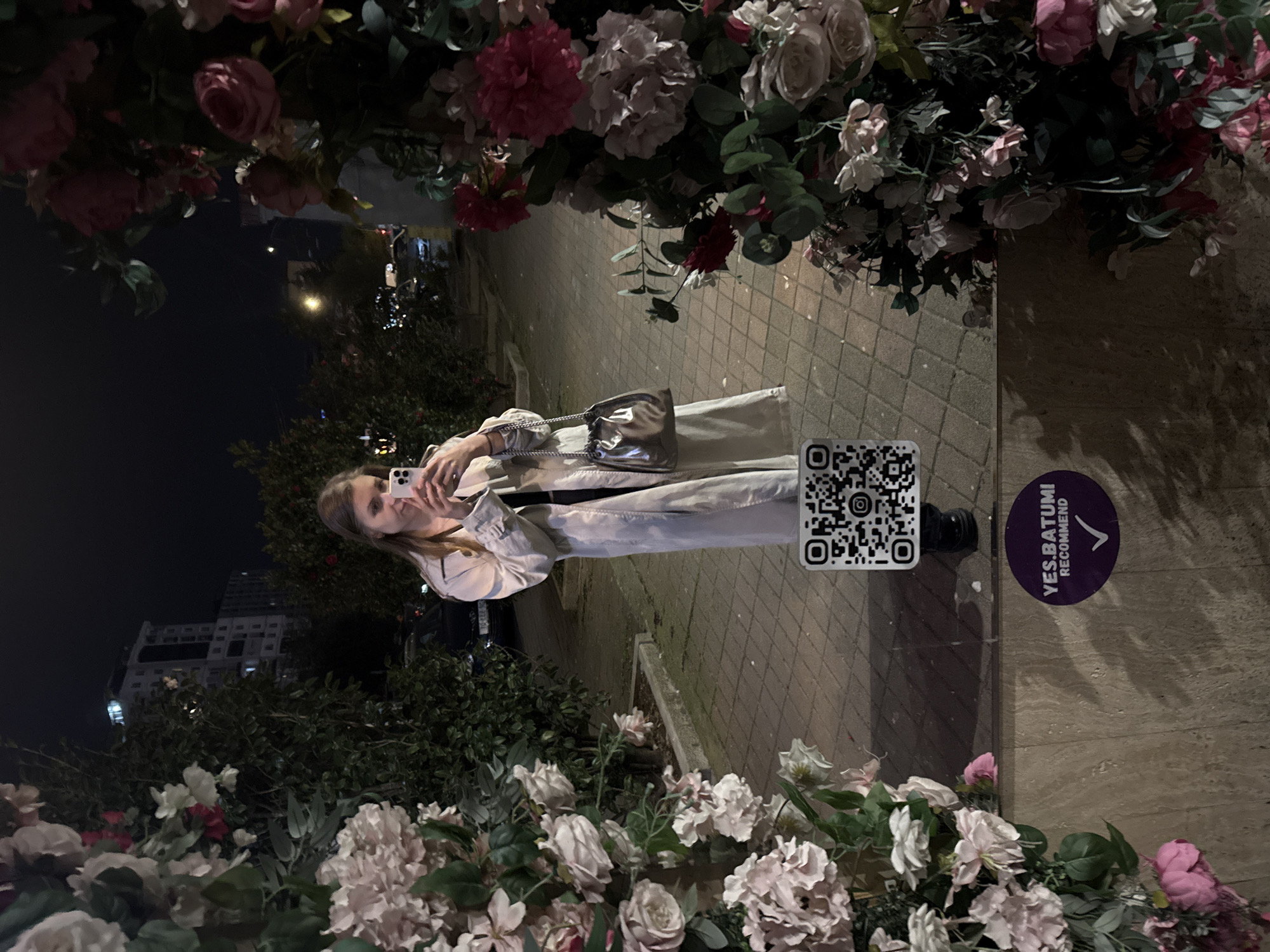 This media file captures a night-time outdoor scene, likely in an urban area of Batumi, Georgia. The main subject is a young woman, seen taking a selfie or mirror picture with a white smartphone. She has light brown hair and is dressed in a light-colored, possibly beige, long coat, carrying a shiny silver handbag with a chain strap. Her expression is neutral as she focuses on her phone.
The woman is framed by a decorative archway or border made of artificial flowers, including pink and white roses and peonies with green foliage. In the background, street lights illuminate a paved walkway, and a tall building is visible in the distance under the dark night sky.
Notable details include a black and white QR code sticker affixed to a surface near the woman's foot. Beside it, a purple circular sticker clearly displays the text "YES.BATUMI" and "RECOMMEND," confirming the location context of Batumi, Georgia. The artificial lighting suggests it's late evening or night, creating a warm glow against the dark environment.