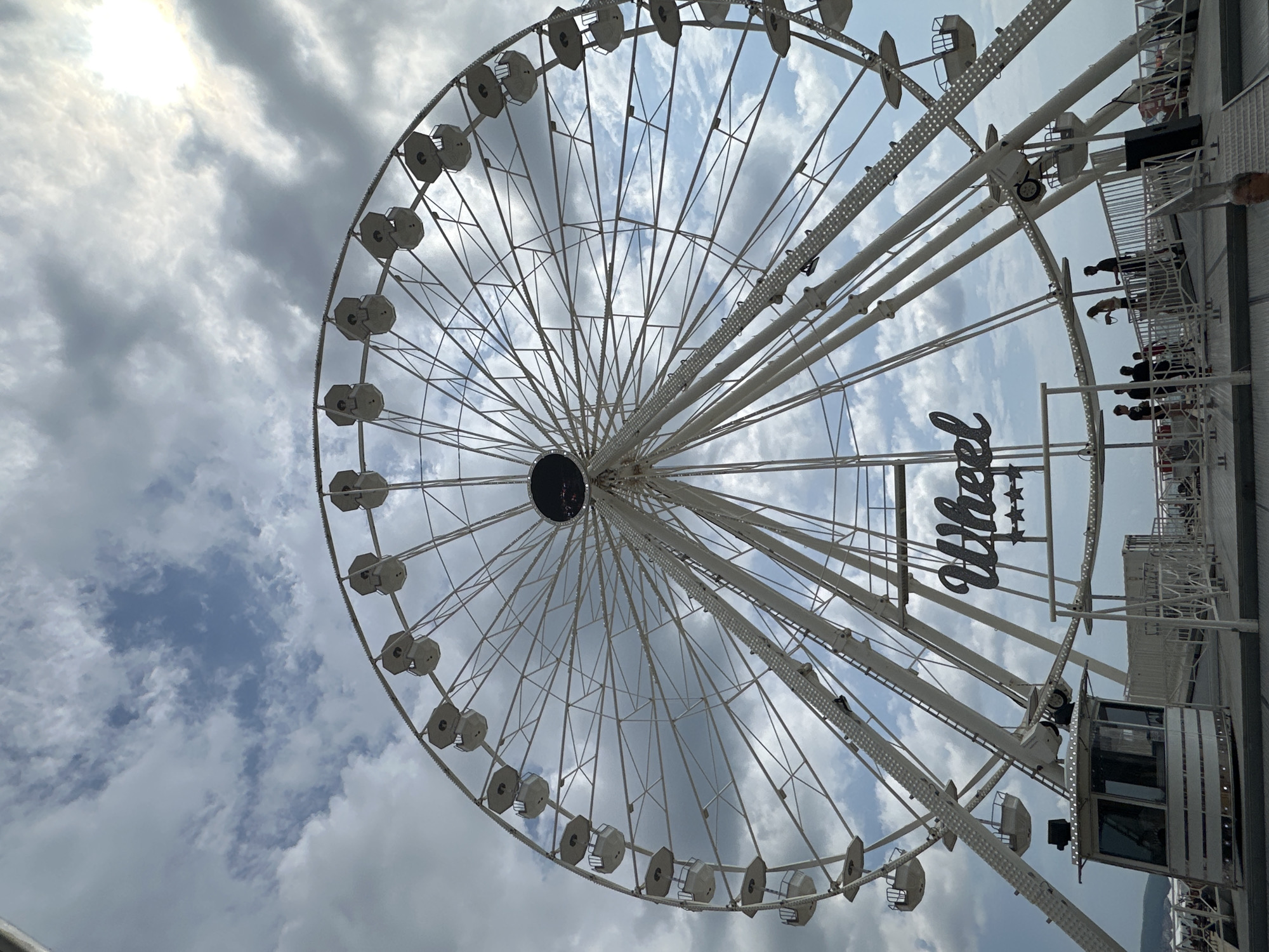 A large white Ferris wheel dominates the frame, set against a sky filled with scattered clouds. The Ferris wheel has numerous white gondolas attached to its rim and a central dark circular structure. Below the Ferris wheel, a sign with "Wheel" written in a stylized font and four stars is visible. There are several people standing on a platform in front of the Ferris wheel, near a small control booth. The scene appears to be an amusement park or a similar recreational area.