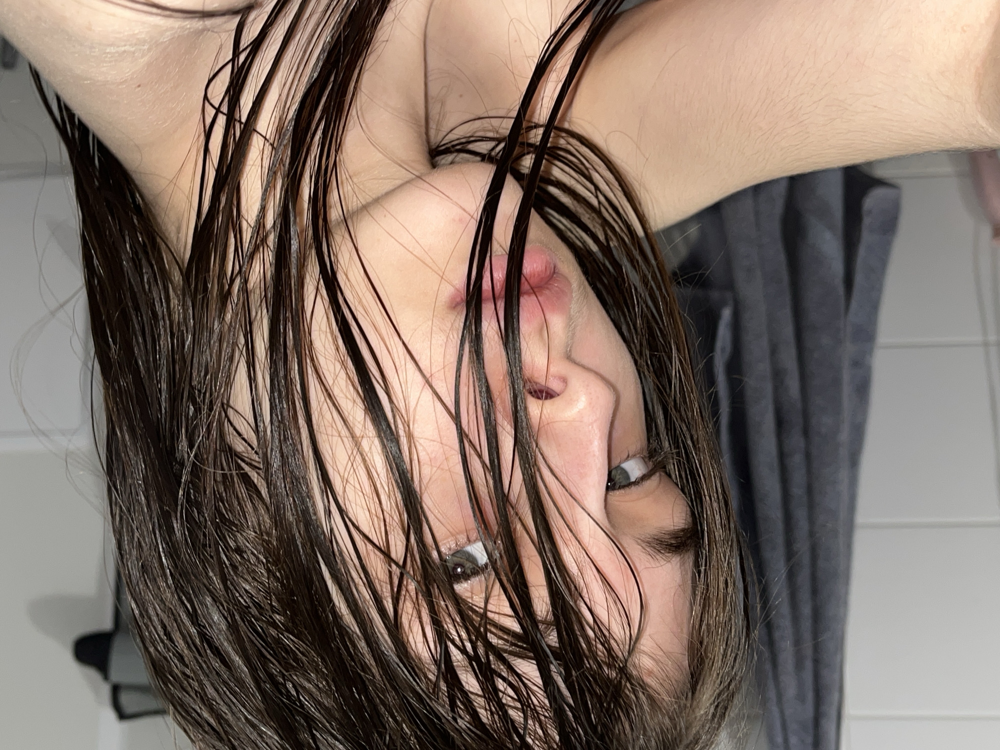 A close-up, upside-down view of a person's face, likely after a shower or bath, with wet, dark hair hanging down. The person's eyes are open and appear to be looking at the camera. The background includes a grey towel and a white tiled wall. The image does not contain sufficient information to identify a location in Saint Petersburg, Russia.