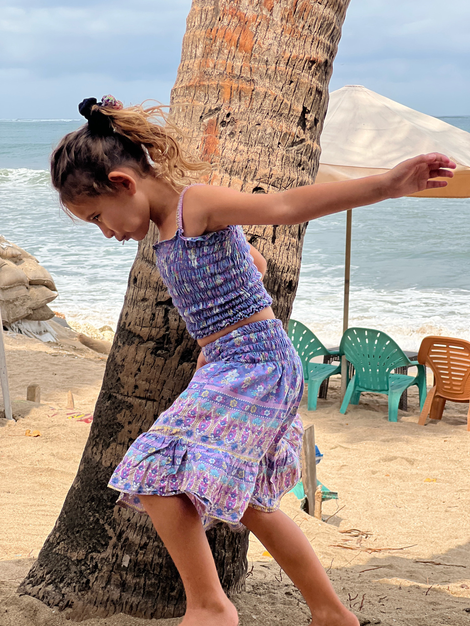 A young girl with brown hair in a ponytail, wearing a purple floral print two-piece outfit, stands on a sandy beach in Kuta, Indonesia. She is leaning against a palm tree, with her right arm extended and her left hand reaching out. The ocean with waves is visible in the background, along with several beach chairs and a large umbrella. The lighting suggests it is daytime, with some overcast sky.