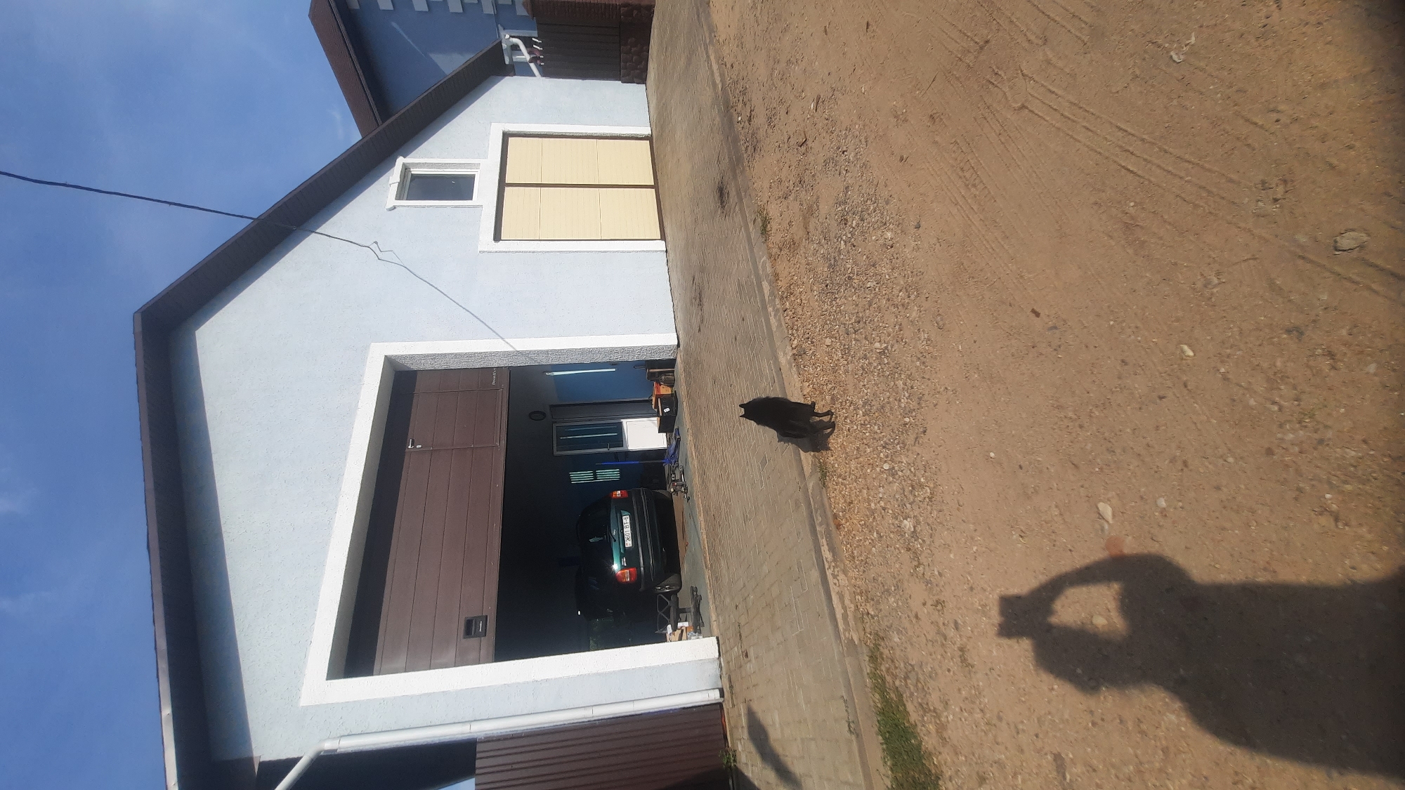 A black dog stands on a paved surface in front of a light blue garage. The garage has a large brown door that is partially open, revealing a dark-colored car inside. To the right of the open garage door, there is a closed yellow garage door and a small window. The garage is a single-story building with a dark brown roof. A shadow of a person holding a phone is visible in the lower left corner of the image, indicating the photographer. The ground in front of the garage is a mixture of paved surface and dirt. The sky is blue with some clouds. The scene appears to be in a residential area, likely in the city of Shklov, Belarus.