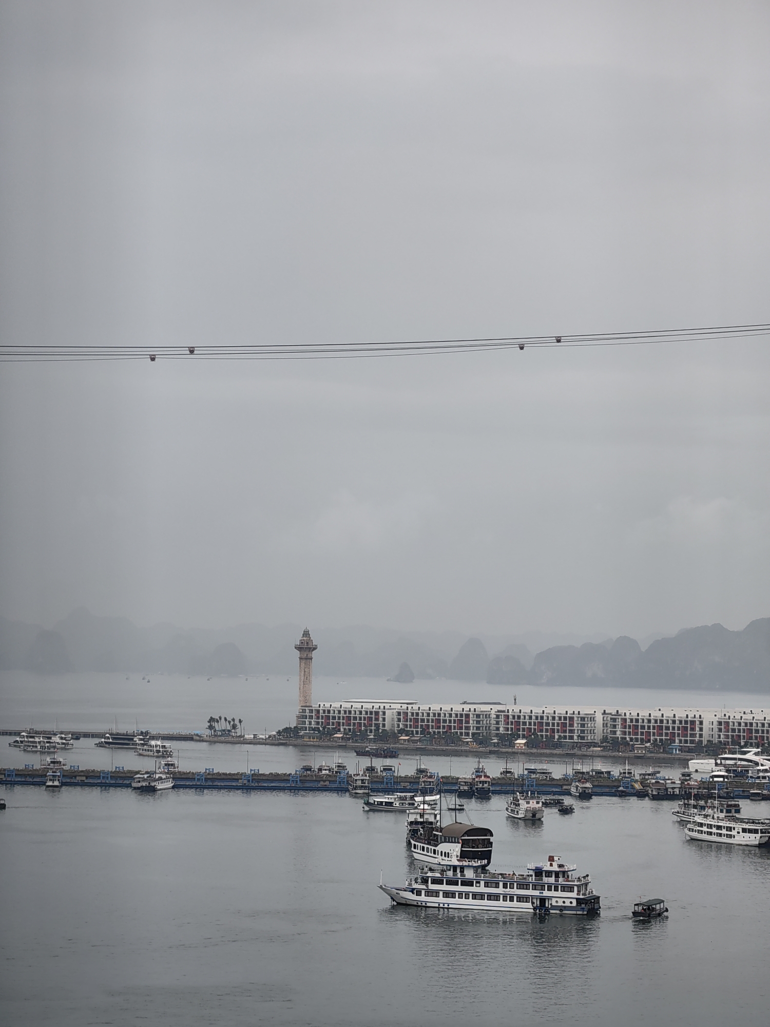 The image shows a harbor in Bai Chay Ward, Vietnam, under an overcast sky. The water is calm and gray. Multiple boats of various sizes are docked or moving within the harbor. A long building, predominantly white with red accents, lines the waterfront. To the left of the building, a tall, light-colored tower stands. In the distance, hazy land formations are visible. A set of wires runs horizontally across the upper portion of the frame.