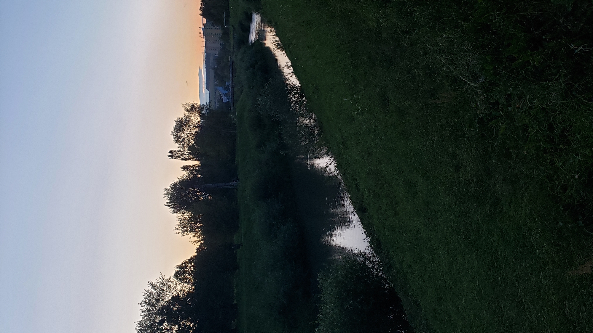 This image depicts a serene riverbank scene at dusk in Prešov, Slovakia. A calm river meanders through lush green grass, reflecting the soft twilight sky. Tall trees line the opposite bank, their silhouettes dark against the fading light. In the distance, the outlines of buildings and a bridge are visible, suggesting the presence of a suburban or urban edge to this natural setting. The overall atmosphere is peaceful and tranquil, characteristic of a quiet evening in Prešov.