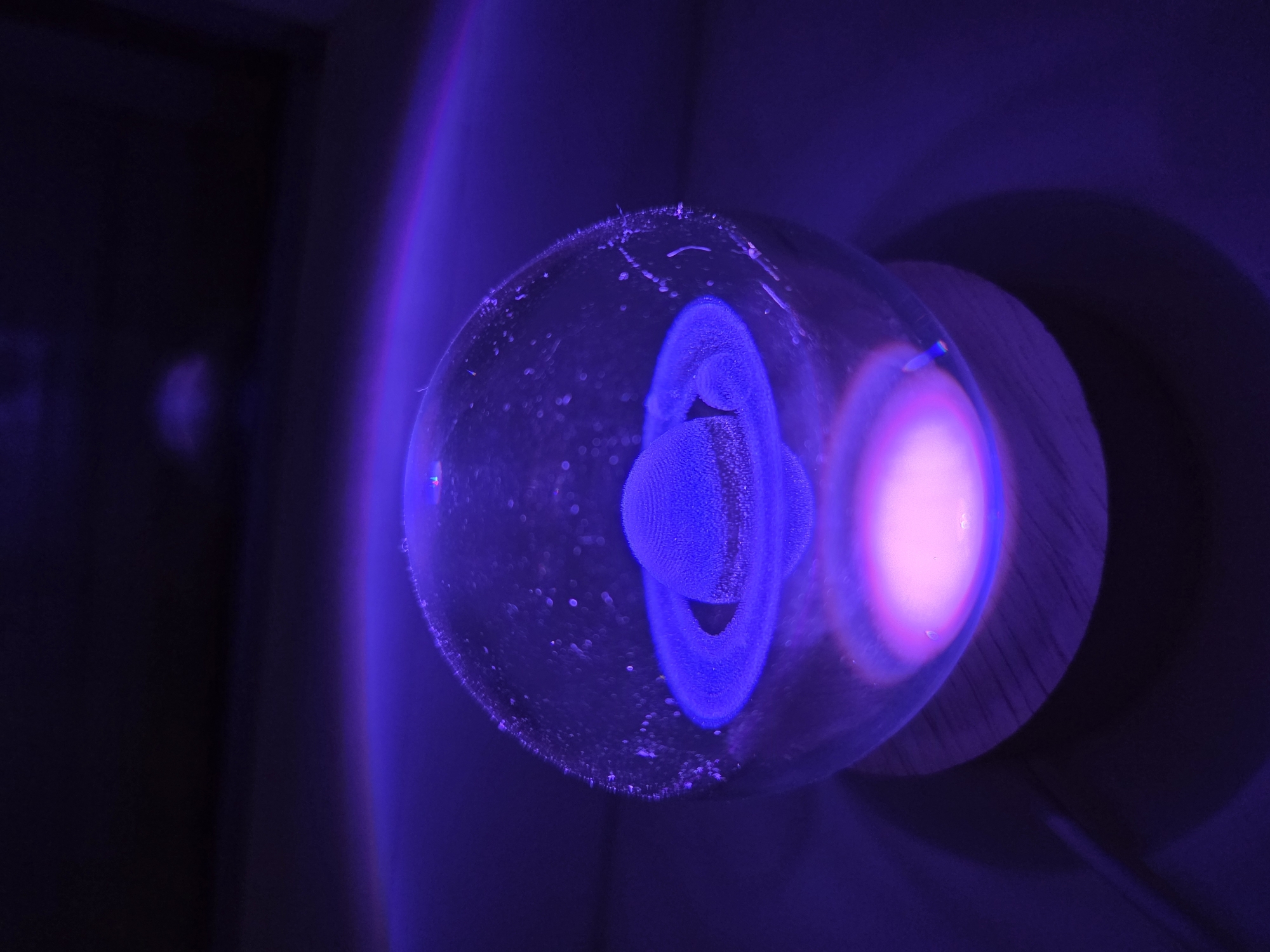 A glass sphere containing a blue, ringed object is illuminated with purple and pink light. The sphere sits atop a wooden base. The background is dark and indistinct.