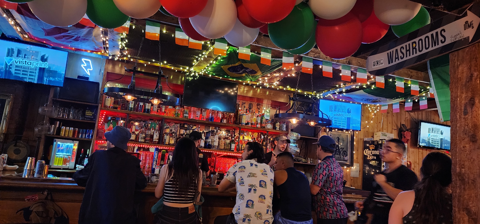 The image shows the interior of a dimly lit bar. A long wooden bar counter is in the foreground, with several patrons seated or standing. Behind the bar is a wall lined with shelves stocked with bottles of alcohol. Red LED lights illuminate the shelves. A large industrial-style chandelier hangs above the bar.
Several screens are mounted on the wall, displaying logos and advertisements. A sign overhead reads "WASHROOMS" with an arrow pointing to the right. The ceiling is decorated with balloons and strings of lights. Small flags, seemingly of Ireland and Brazil, are strung across the ceiling.
In the center of the image, a bartender is serving patrons. One patron, wearing a blue hat, is looking at the bar. Another patron in a black shirt is talking to the bartender. On the right, a person in a colorful patterned shirt is standing. Further to the right, a person in a black shirt and glasses is talking to another person whose back is to the camera.
The overall atmosphere appears casual and lively, typical of a bar or pub. The location is identified as Unknown, Unknown.