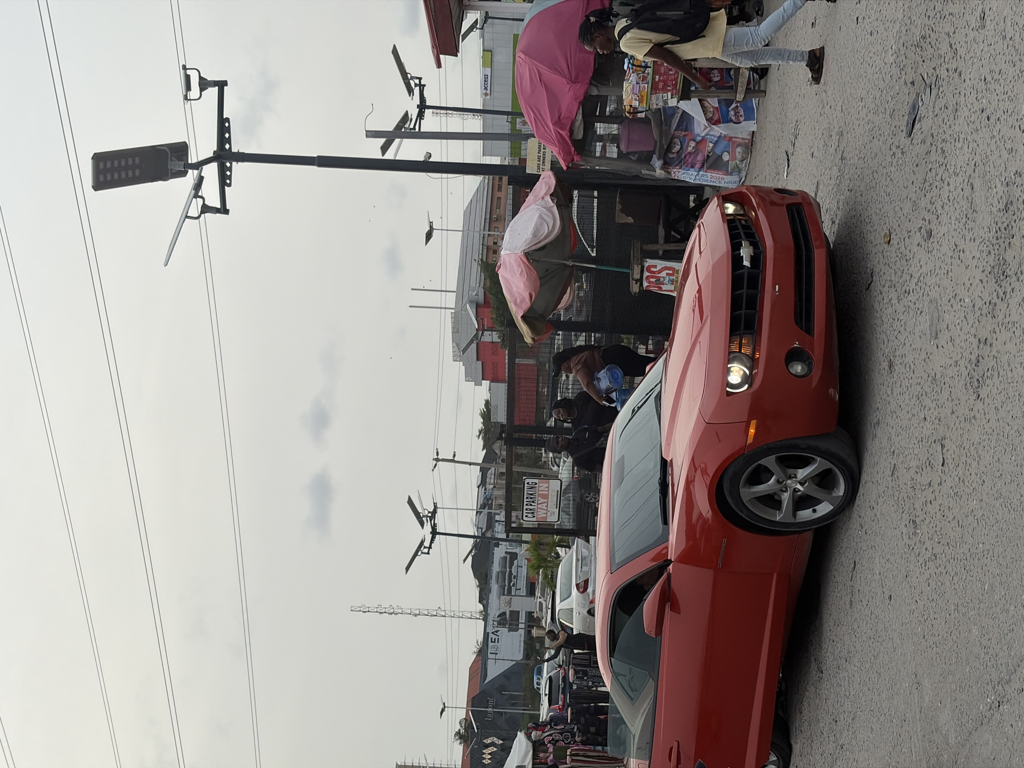 This media file captures a daytime street scene in Igboefon, Nigeria, characterized by urban activity and commercial presence under an overcast sky.
In the foreground, a prominent red Chevrolet Camaro is seen from the front-right side, positioned on what appears to be a paved road or parking area. Its headlights are illuminated. To the left of the car, along a sidewalk or shoulder, street vendors operate under two large, colorful umbrellas, one pink and another pink and brown. Various items of merchandise are visible near these stalls, and a "POS" (Point of Sale) sign is seen on the ground.
Several individuals are present: a person in a yellow top and light trousers with a backpack is walking past the vendor stalls, and another person is bending down near the front of the red car. In the background, numerous buildings line the street, featuring various signs. Overhead, a dense network of power lines crisscrosses the sky, and a tall street light pole equipped with solar panels stands on the left. A faint construction crane is visible in the distant, cloudy sky.
Visible text in the image includes a white sign reading "CAR PARKING WAY IN" and "CARS ARE PARKE AT OWNERS RI" (likely "RISK"). Building signs display "access," "lagom," and "SEAT." A poster or banner near the vendors advertises an event, showing "EXT. PRAYERS 2026 CONFERENCE NIGE" (likely "NIGERIA"). The scene depicts a bustling urban environment with ongoing pedestrian and commercial activity.