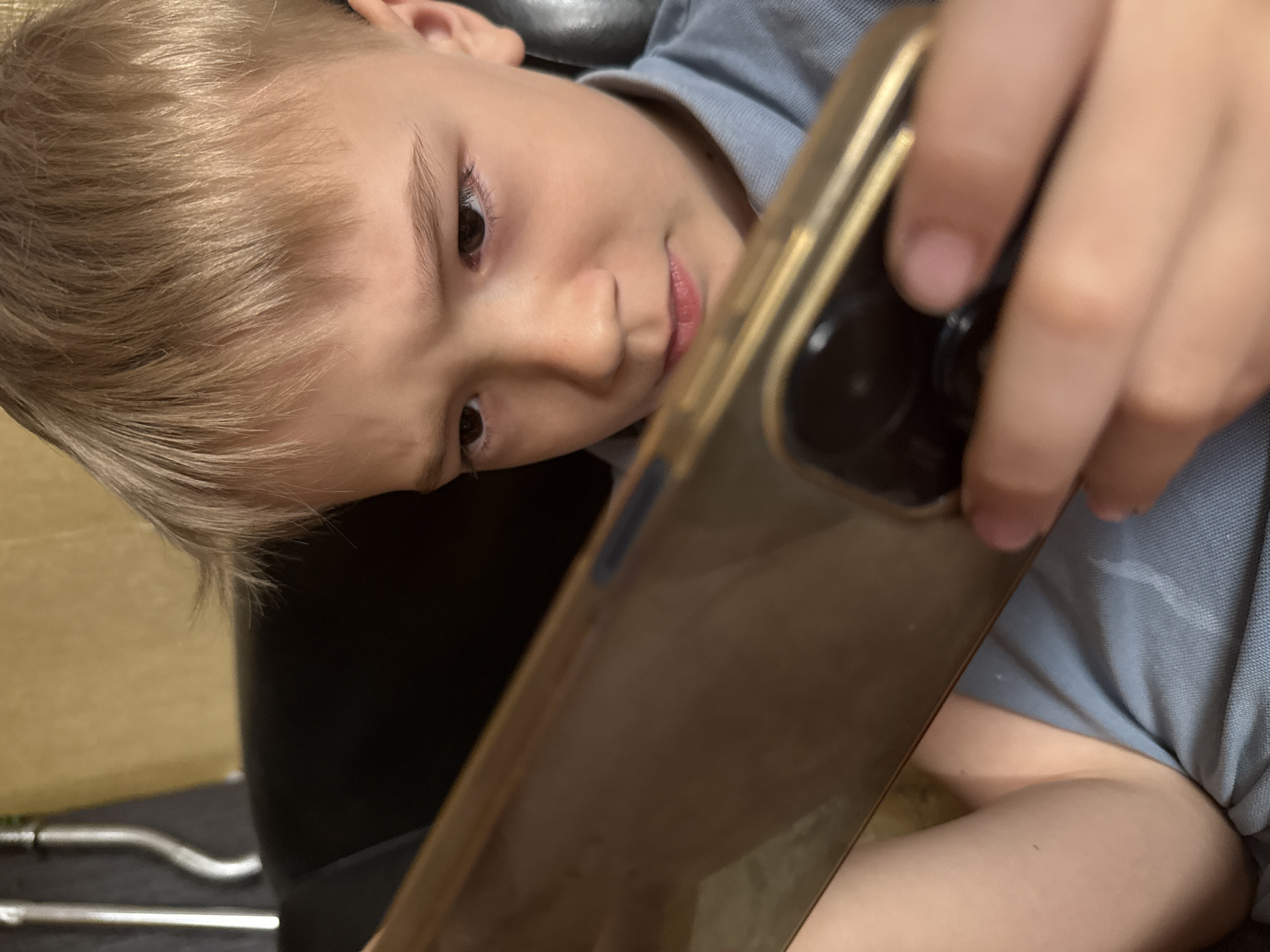 A child with light blonde hair, wearing a light blue shirt, is holding a gold-colored smartphone. The child is looking down at the phone, holding it in front of them with one hand. The background shows a dark-colored surface with a glimpse of what appears to be a metal object or part of furniture. The image is taken at an angle, tilting the subject. No information about Dmitrov, Russia, is discernible from the image.