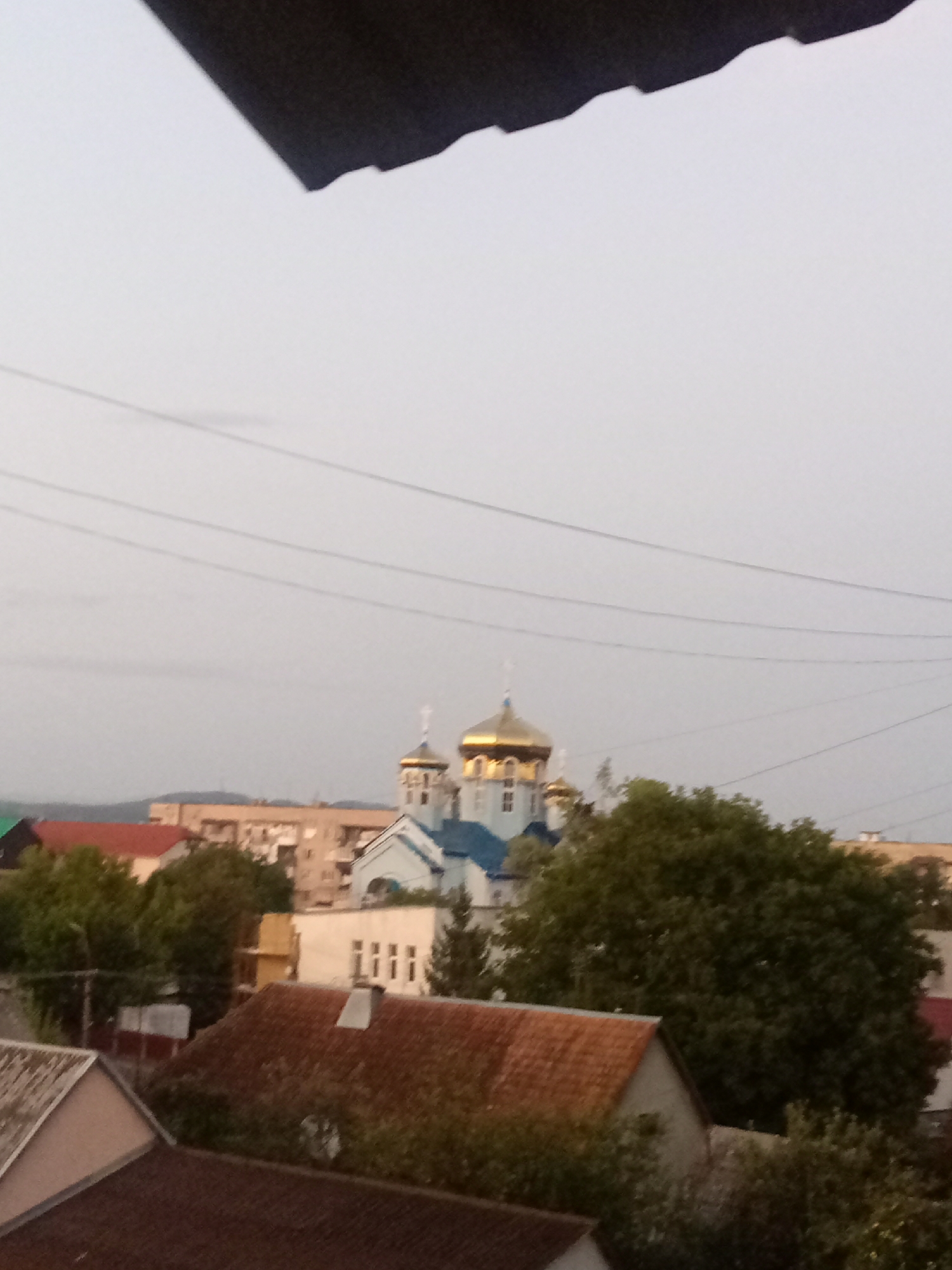 The image captures a view of a city with multiple buildings and lush greenery, under an overcast sky. In the mid-ground, a prominent blue and white church with golden domes stands out, featuring architectural details like arched windows and a cross on top. Several other buildings, varying in size and color, are visible behind and around the church, some with red tiled roofs. A large, dense green tree dominates the right side of the frame, partially obscuring the church. In the foreground, rooftops of residential buildings are visible, one with a reddish-brown tiled roof and a chimney. Power lines stretch horizontally across the upper portion of the image. The overall scene suggests an urban environment in Tiachiv, Ukraine, likely during daylight hours given the illumination of the buildings and landscape.