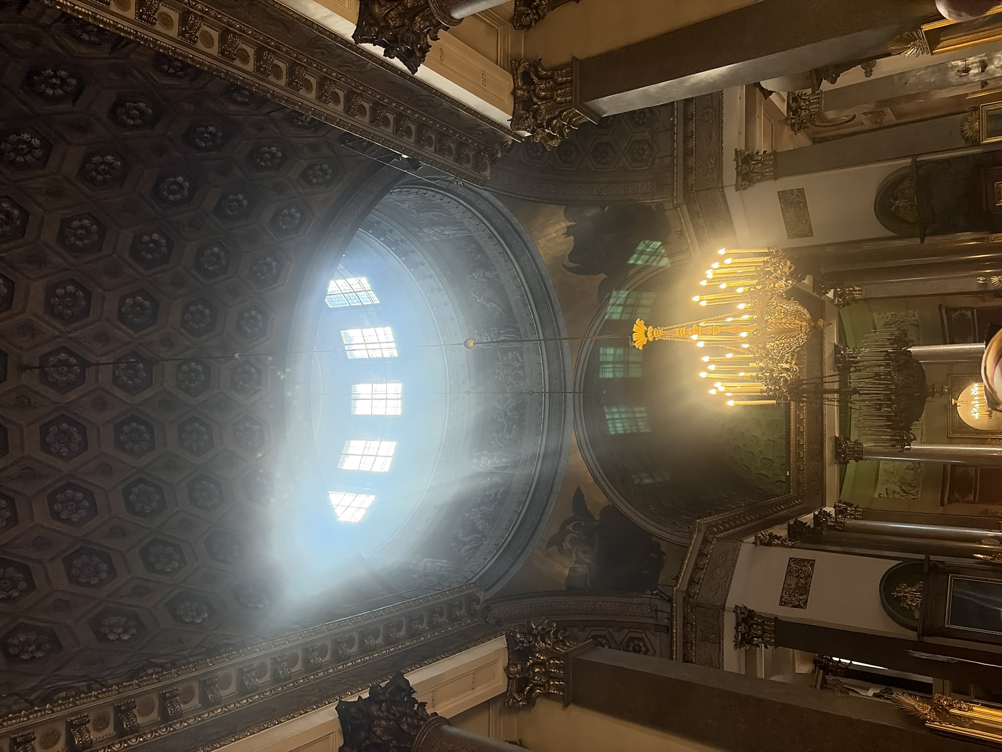 The image shows the interior of a grand cathedral, with a high, ornate ceiling adorned with hexagonal patterns and floral motifs. Sunlight streams dramatically through a large, round oculus in the dome, creating visible beams of light that illuminate dust particles in the air. The dome itself features painted murals depicting figures and scenes. A large, ornate chandelier with numerous lit candles hangs prominently in the foreground, casting a warm glow. Several tall, classical columns with decorative capitals support the structure. The overall impression is one of awe-inspiring architecture and a dramatic play of light and shadow. The location is identified as Unknown, Unknown.
