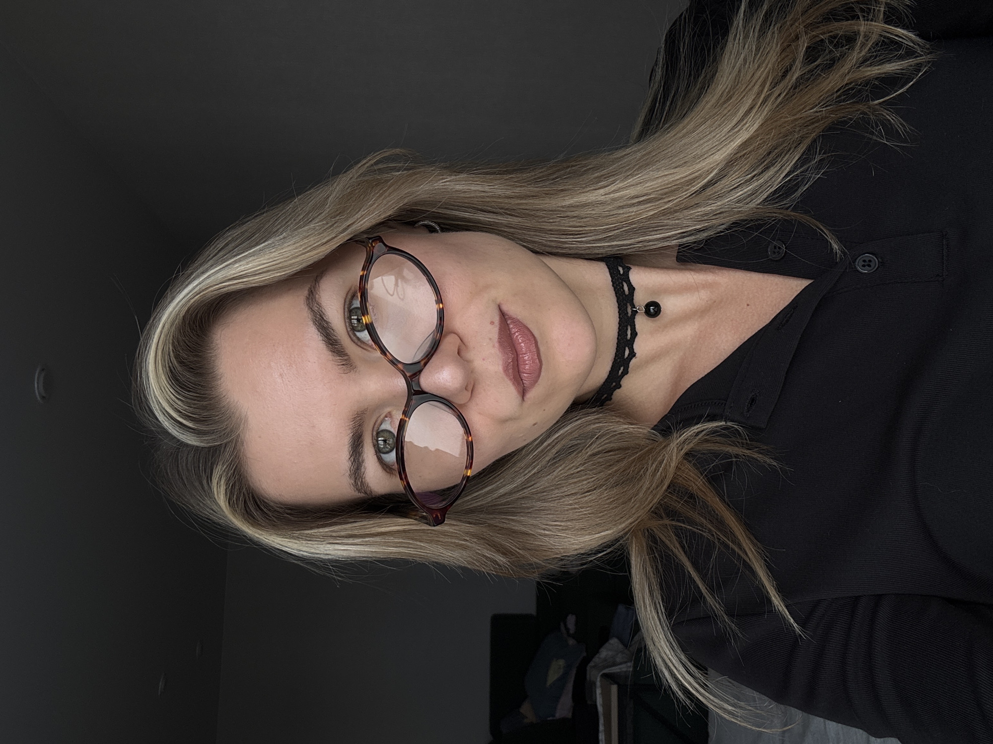 A woman with blonde hair and glasses is shown in a close-up shot. She is wearing a black shirt and a black choker. The background is a dark gray color. The location is Unknown, Unknown.