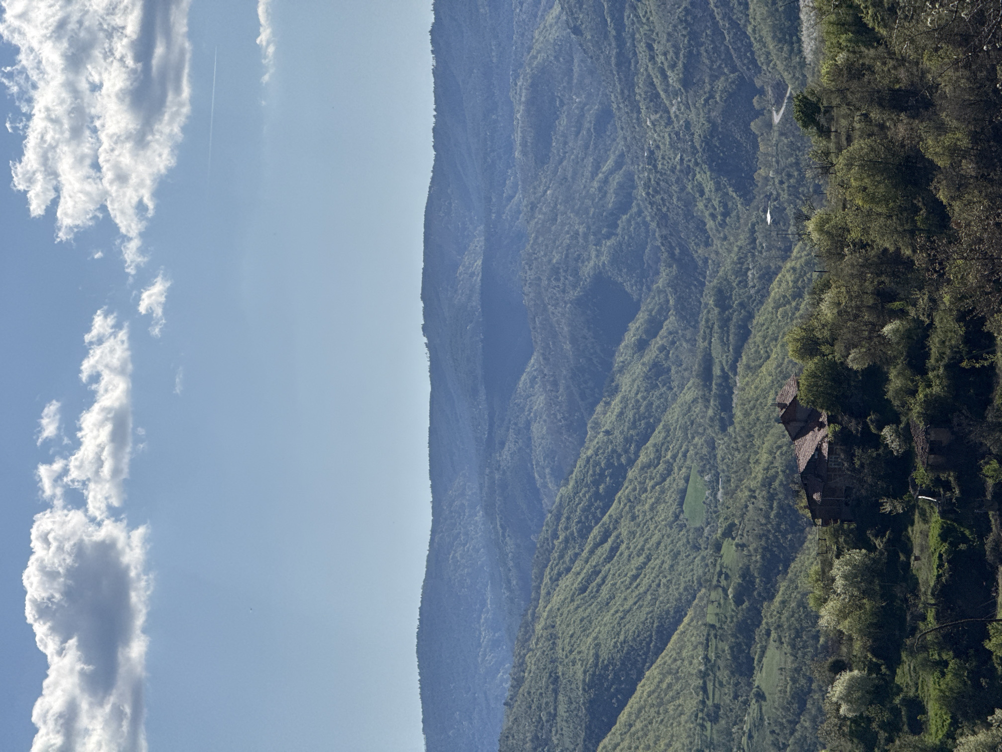 The image shows a landscape, likely in a mountainous region, with a clear sky containing some clouds and a contrail in the upper left. Mountains covered in dense green vegetation fill the right side of the frame. A prominent ridge line delineates the mountains from the sky. On the right side of the image, nestled amidst the vegetation, is a building or structure with a multi-tiered roof. The building appears to be located on a steep slope. The location is Tsarvishte, Bulgaria.